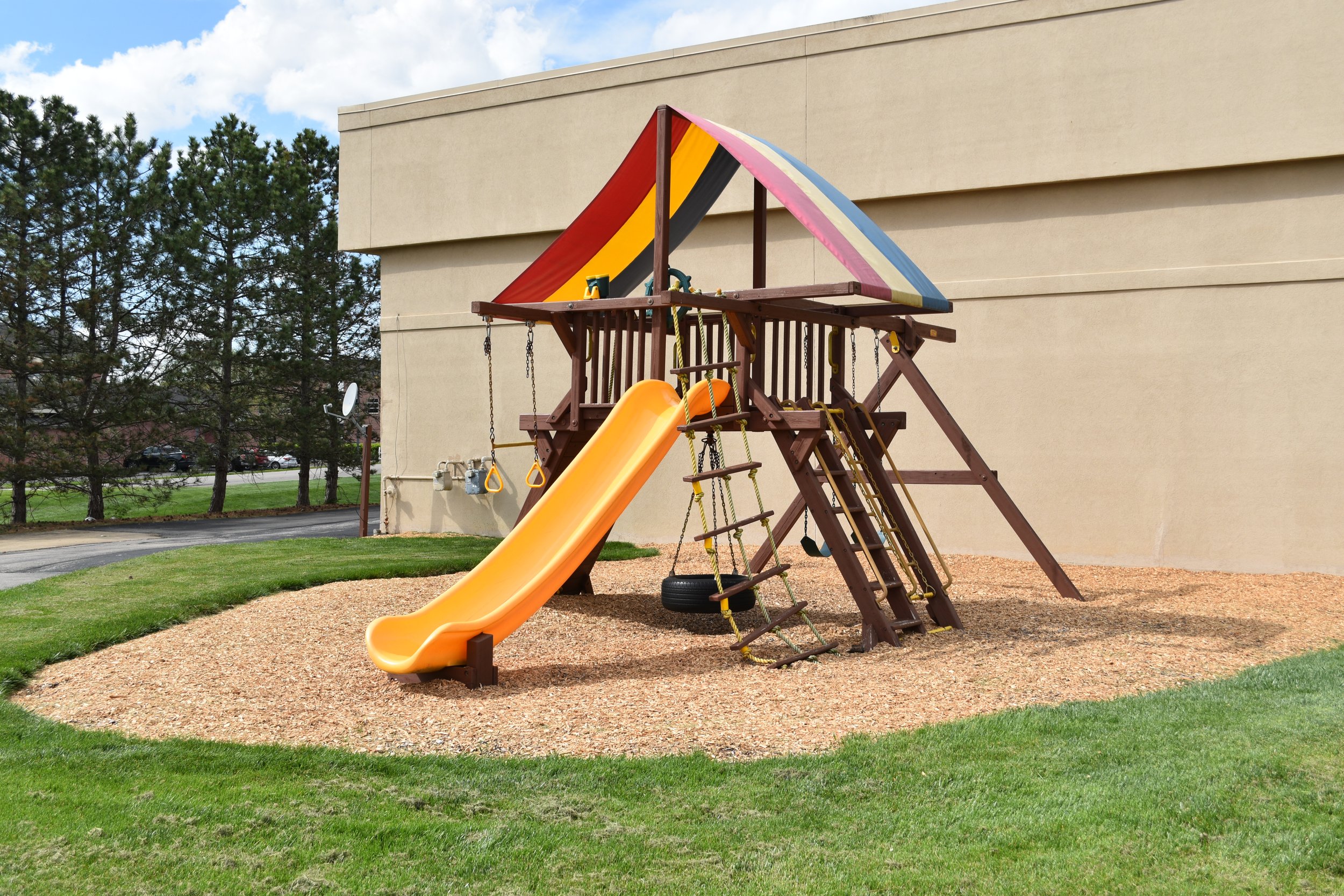 A wooden playground structure with a multicolored canopy, orange slide, swings, and climbing features, located on a bed of wood chips beside a beige building, with trees and parked cars in the background.