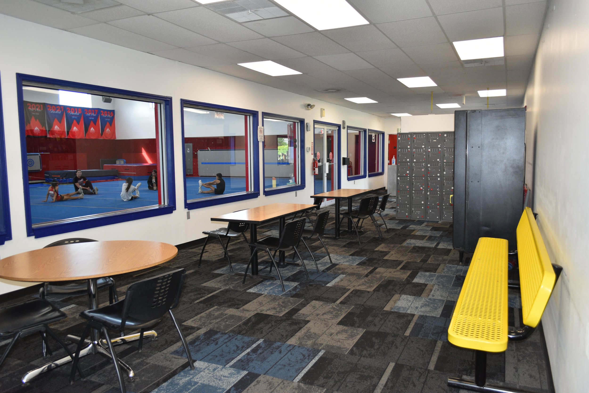 View of an indoor gym with multiple children stretching and practicing on blue mats seen through large windows. The room has a white ceiling with fluorescent lights, lockers on the right, and a yellow bench along the right wall. Tables and chairs are