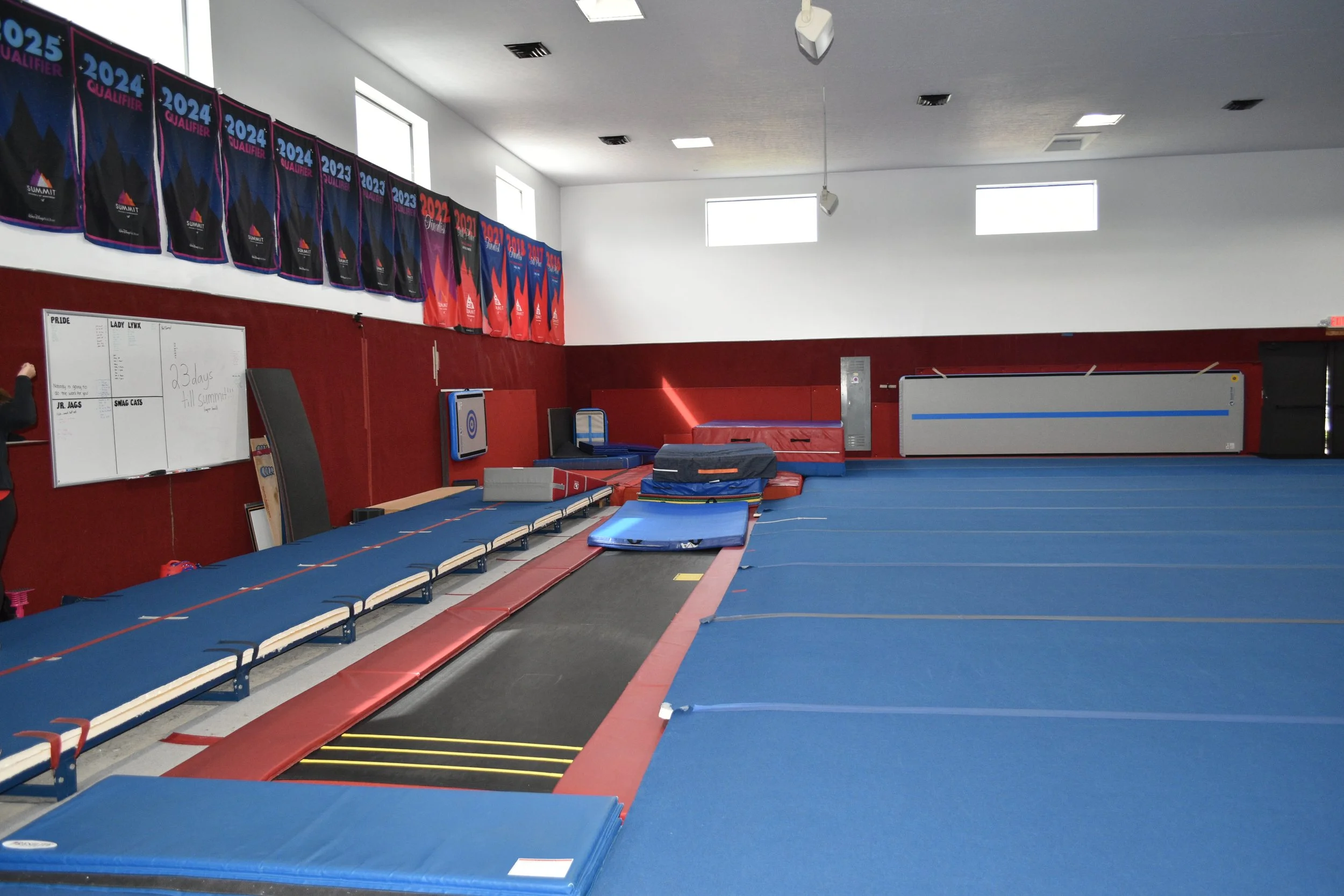 An indoor gymnastics training facility with blue mats, red padding, and various equipment like a trampoline, balance beams, and padded areas. Banners with years and events line the upper wall, and a whiteboard with notes is visible on the wall.