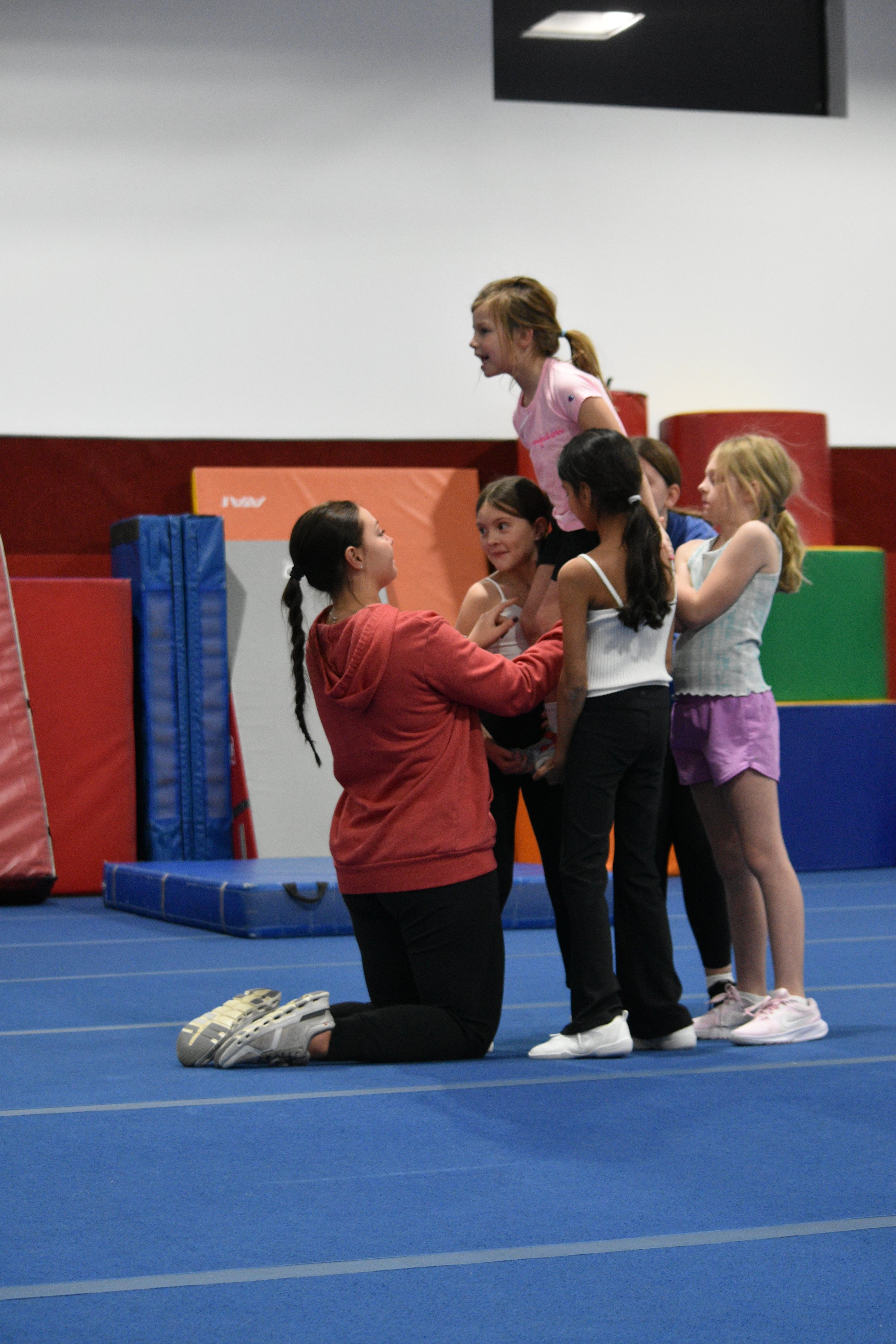 A group of young girls participating in a cheerleading or gymnastics practice in an indoor gymnasium with blue mats, padded walls, and various gym equipment.