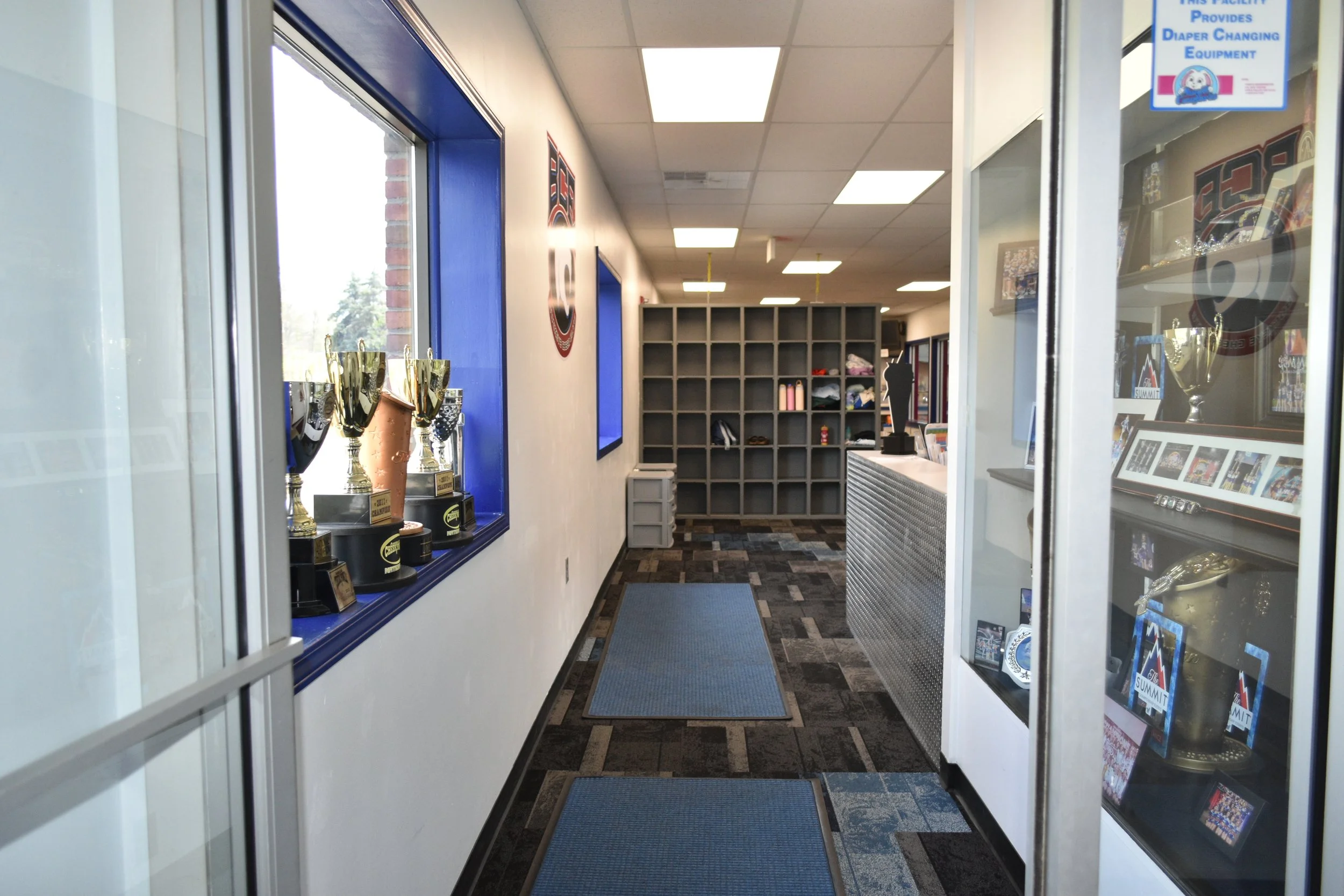 A hallway view inside a facility with trophy display cases on the left and shelves with various items on the right. The floor has cushioned mats, and the ceiling has fluorescent lights.