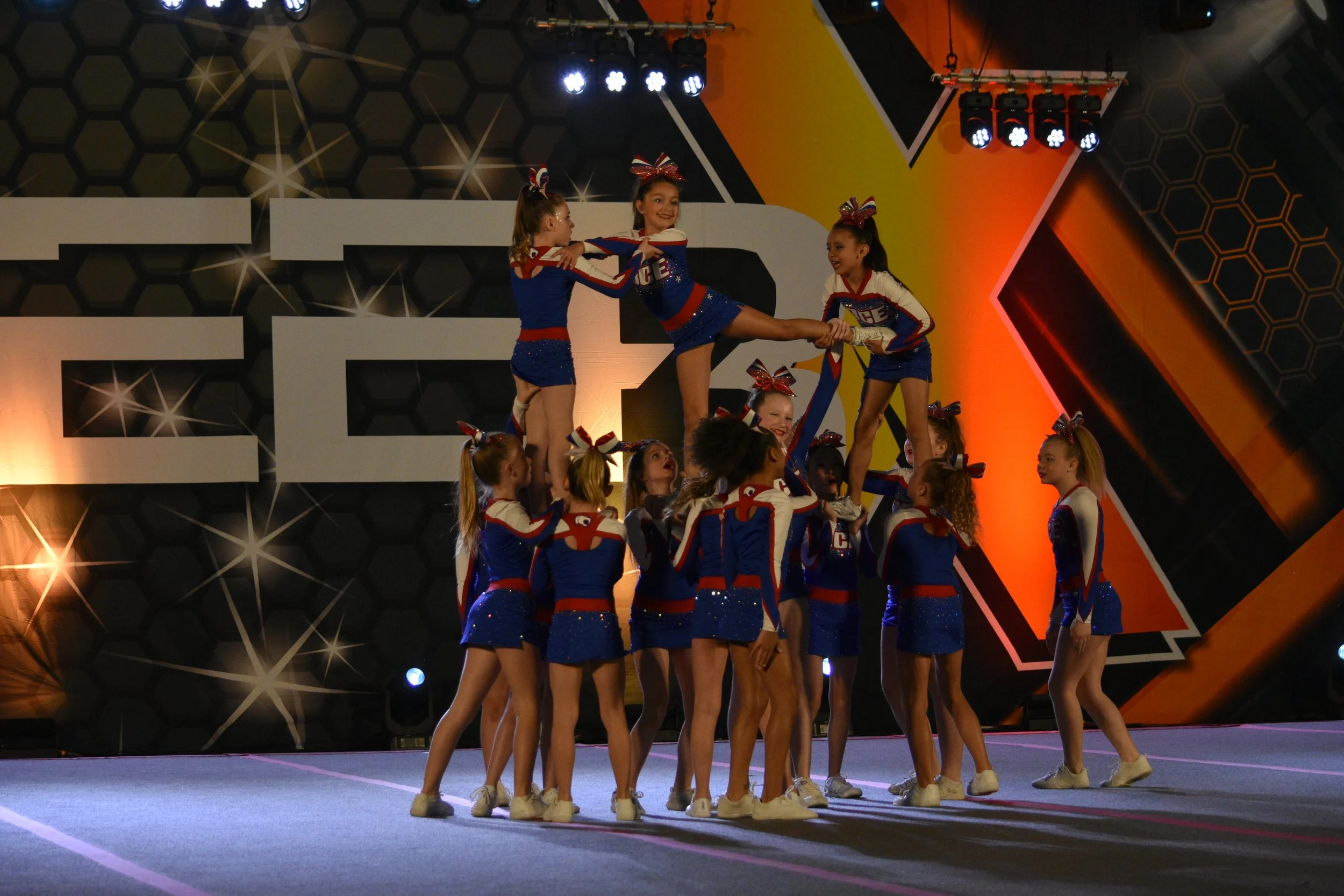 Cheerleaders performing a routine on a stage with a dark, honeycomb-patterned backdrop, dressed in matching red, white, and blue uniforms with bows in their hair, under bright stage lights.