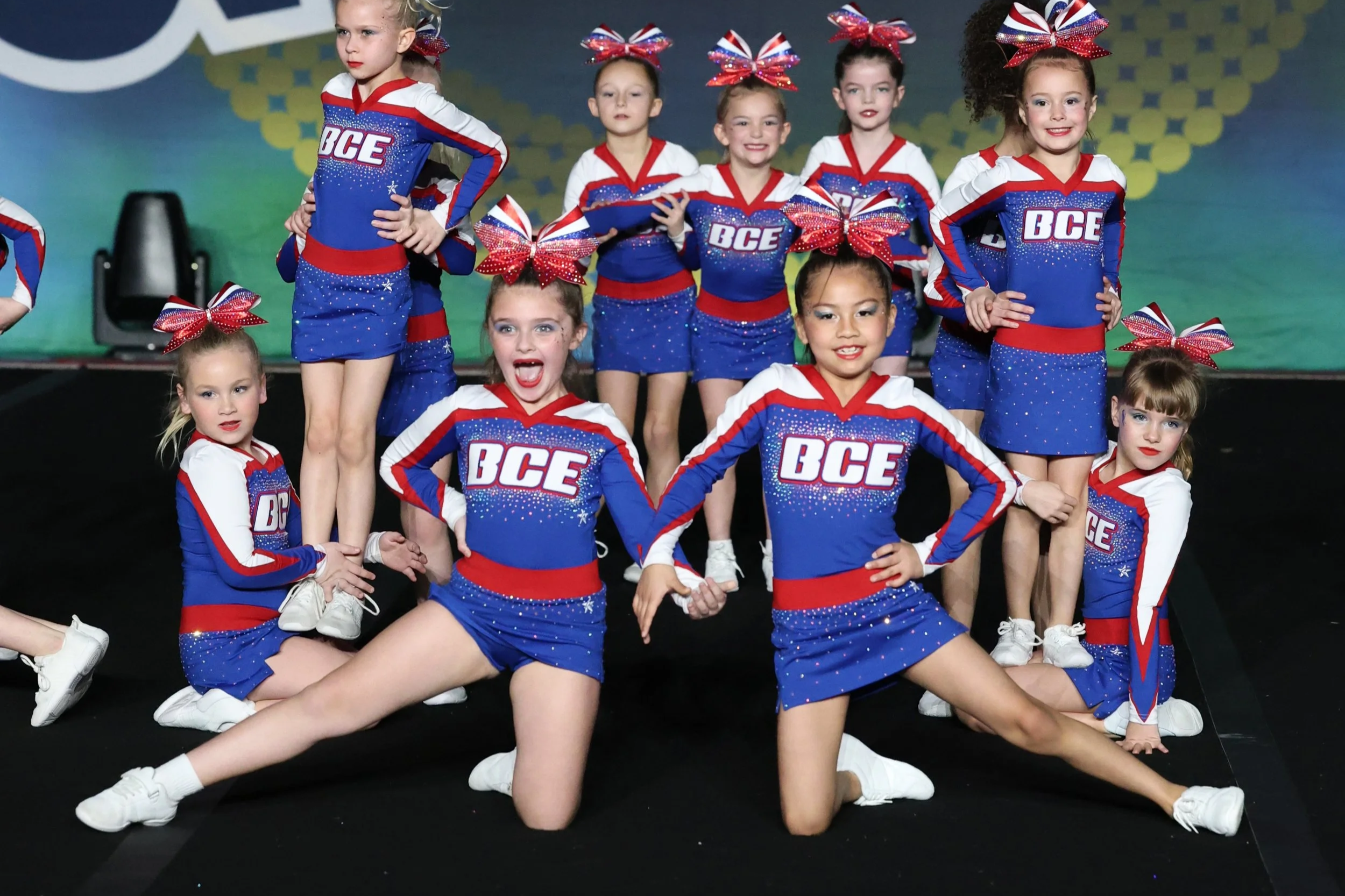 A group of young girls dressed in cheerleading uniforms with red, blue, and white colors, and large red, blue, and white bows in their hair, posing on a stage during a cheerleading performance.