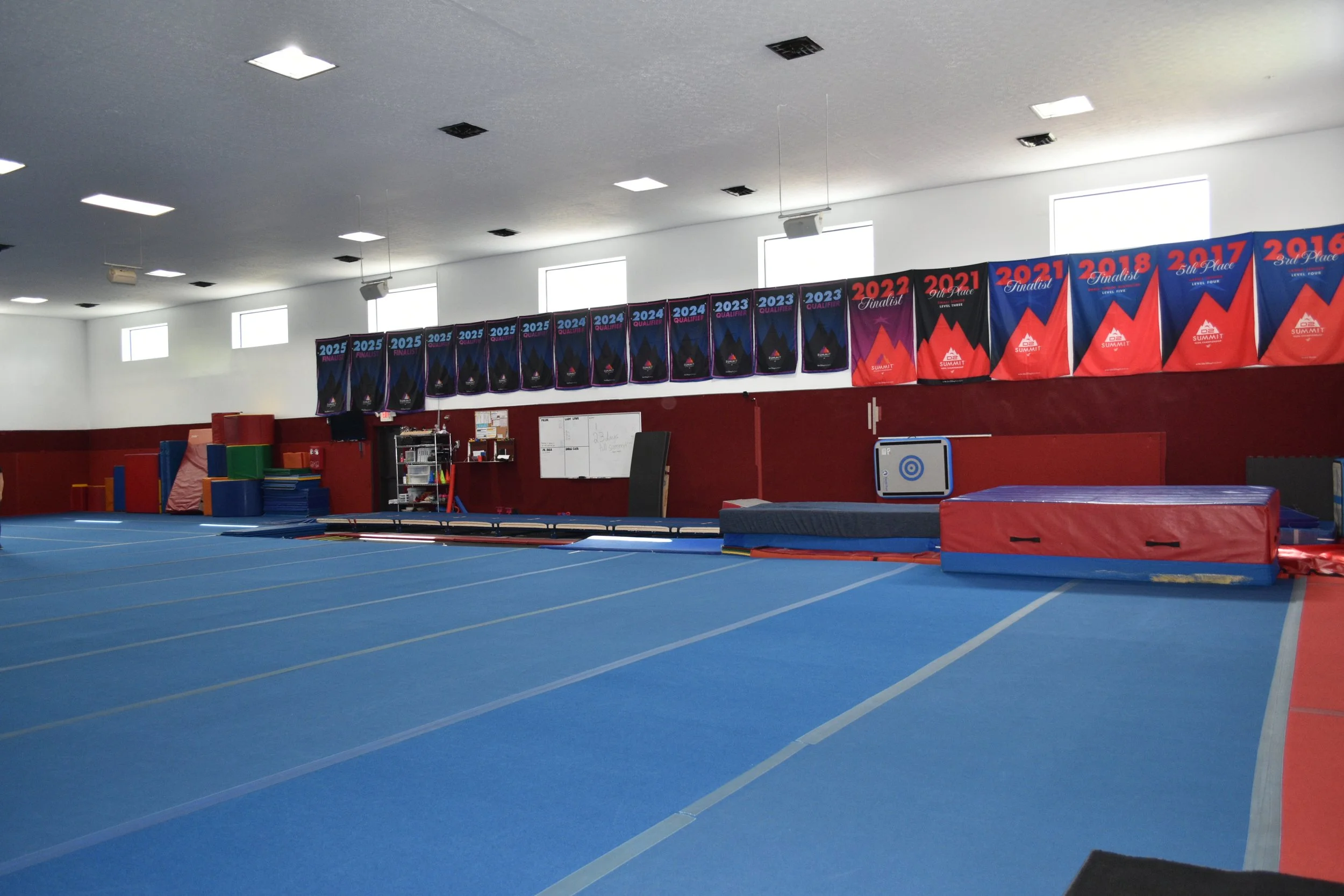Interior of a gymnastics training facility with blue mats, red and blue banners on the wall displaying competition placements from 2017 to 2025, and equipment like mats and foam blocks.
