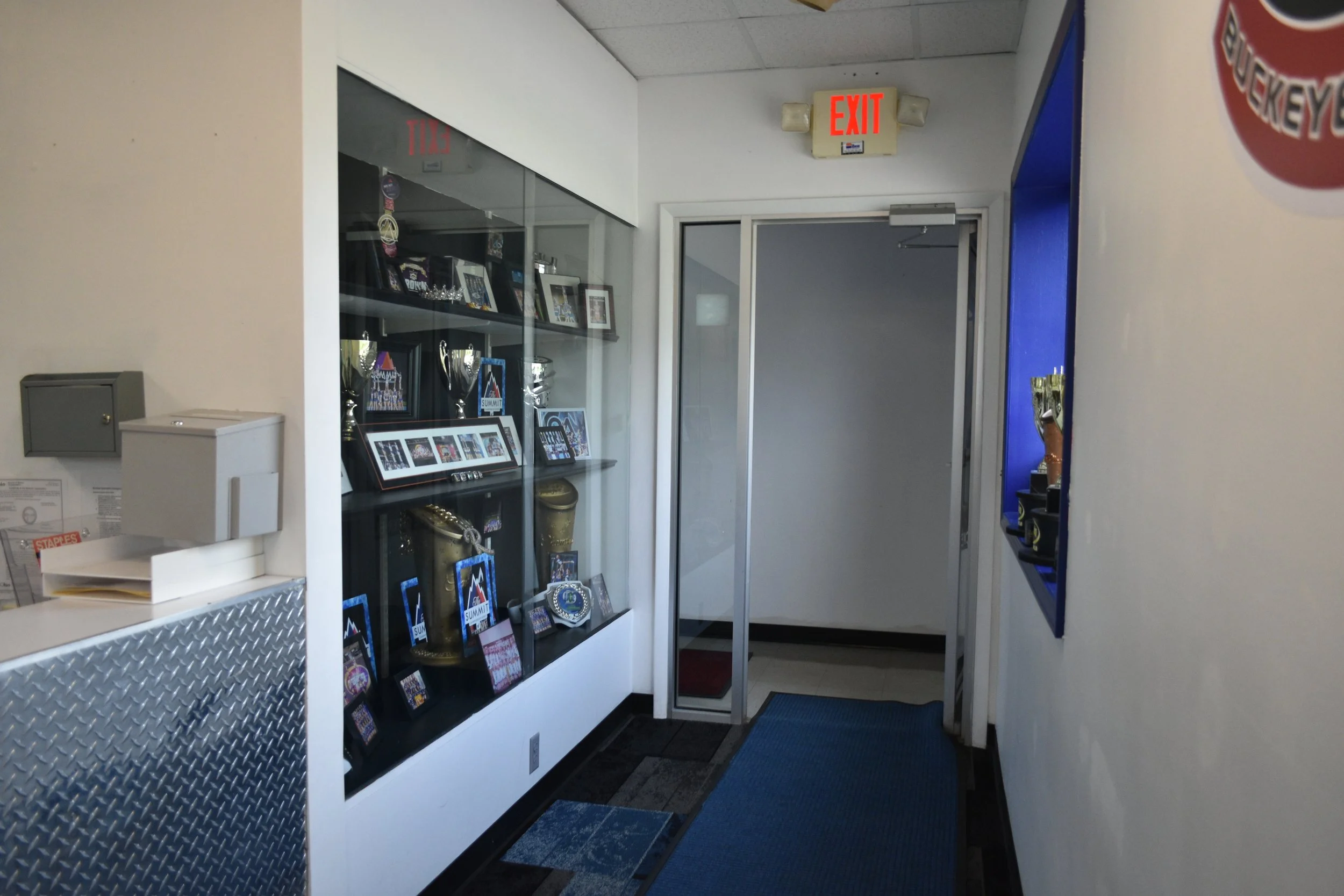 Indoor hallway with a glass display case containing trophies and framed photos, a closed metal door with a glass window, and a white wall with an illuminated red 'EXIT' sign above the door.