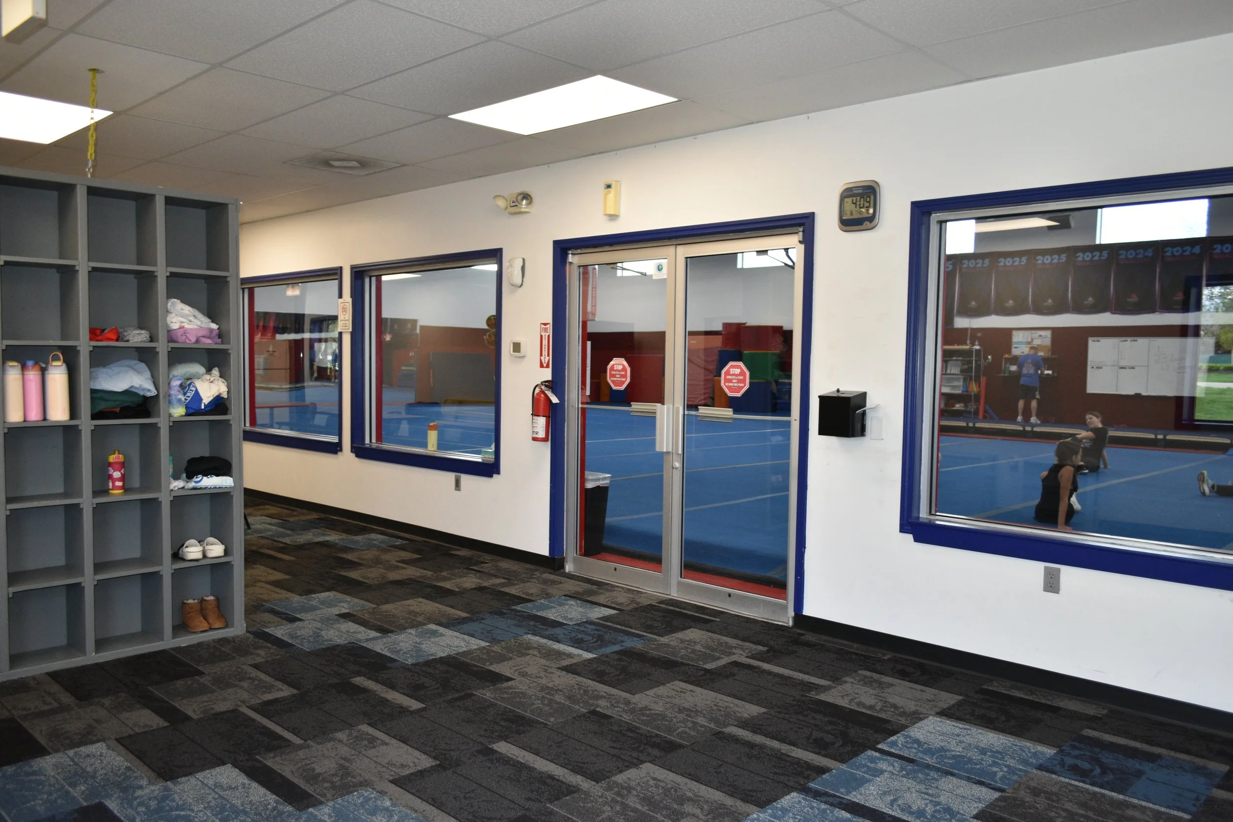 View of an indoor gym facility with a blue court visible through glass doors and windows, and a person exercising inside. The area has a checkered dark gray and blue carpeted floor, a gray storage shelf with folded clothes and water bottles, and seve