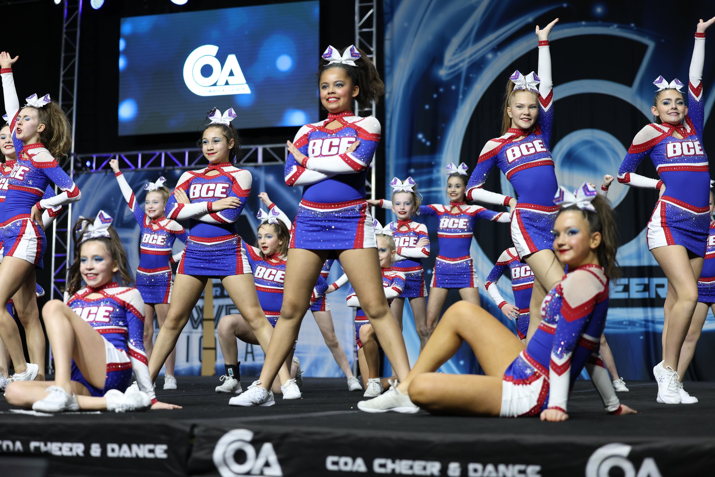 Cheerleaders performing a split on stage during a cheer and dance competition, dressed in red, white, and blue uniforms with big bows in their hair.