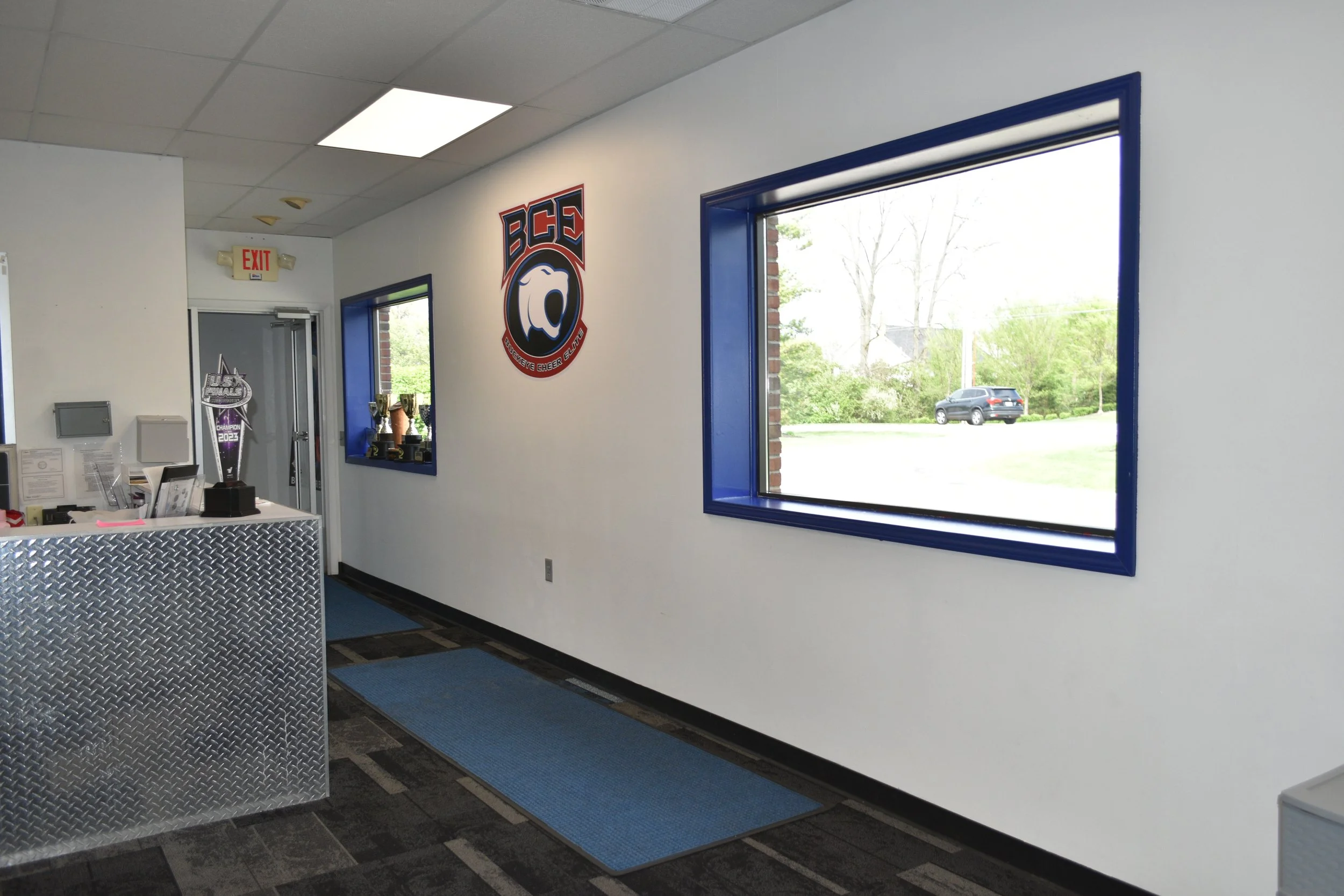 Interior of a cheerleading facility with NBC cheerleading trophies on display, a large window showing a parked SUV outside, and a reception desk with chevron patterned surface.