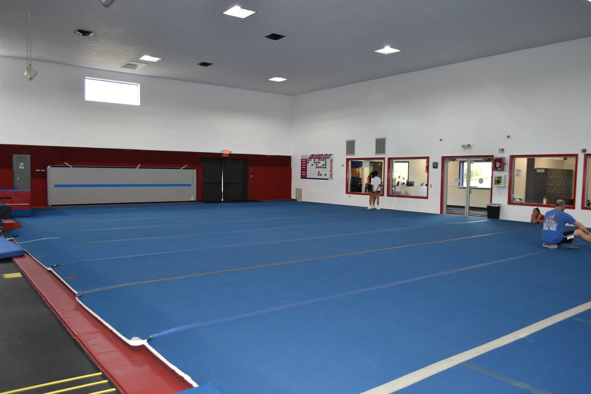 Interior of a gym with blue mats on the floor, a red and white border, some gym equipment on the side, and a person sitting on the floor. There are windows and a reception area visible through a glass window.