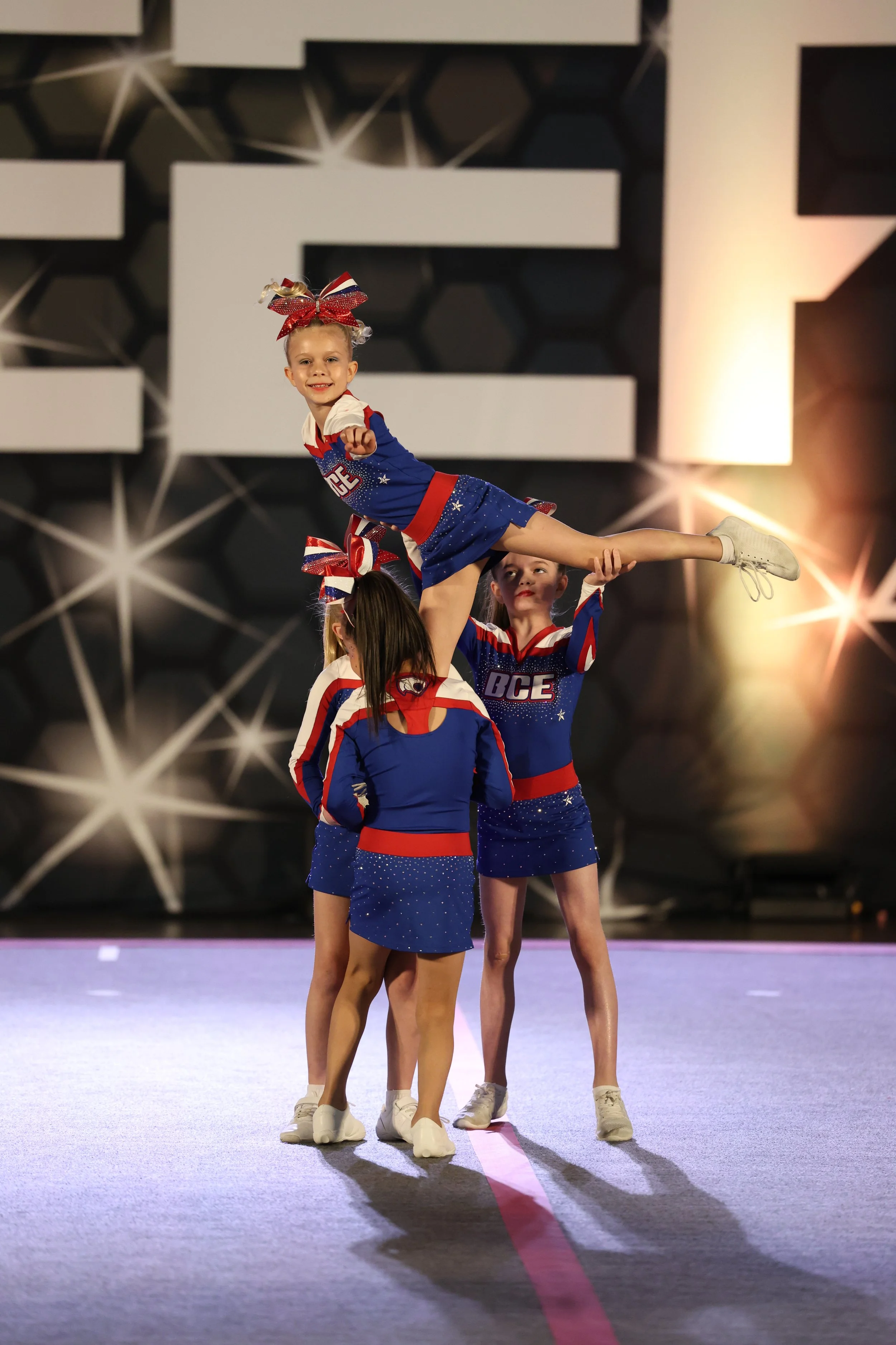 Children cheerleaders in red, white, and blue cheerleading uniforms, performing a routine on a stage with a large, geometric background and bright overhead lighting.
