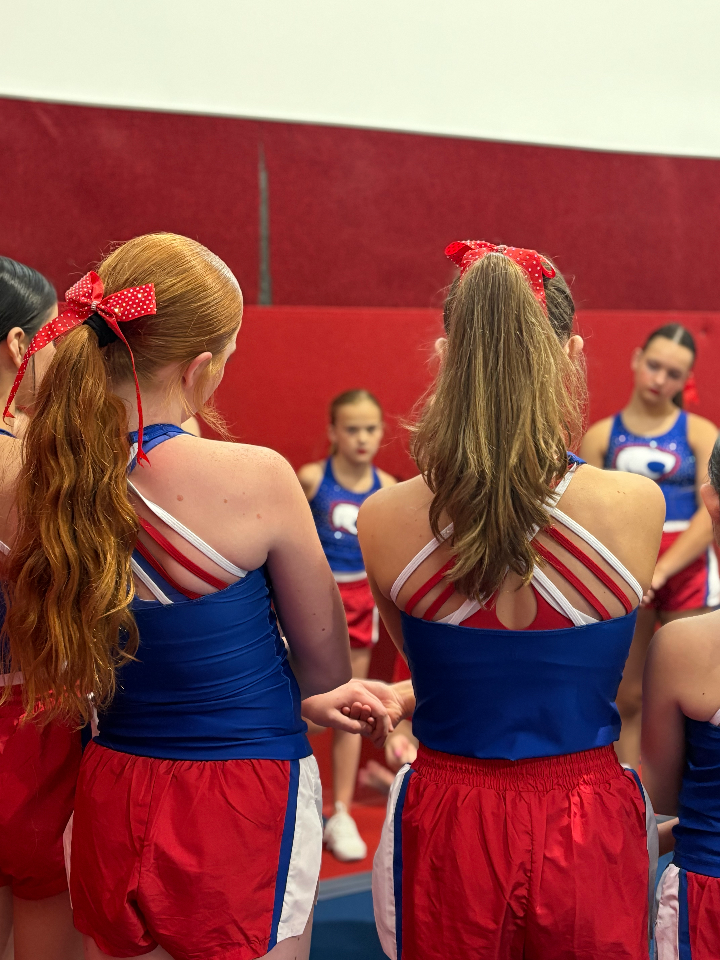 Cheerleaders wearing red, white, and blue uniforms with red polka dot bows in their hair, standing and talking together indoors with a red and white wall behind them.