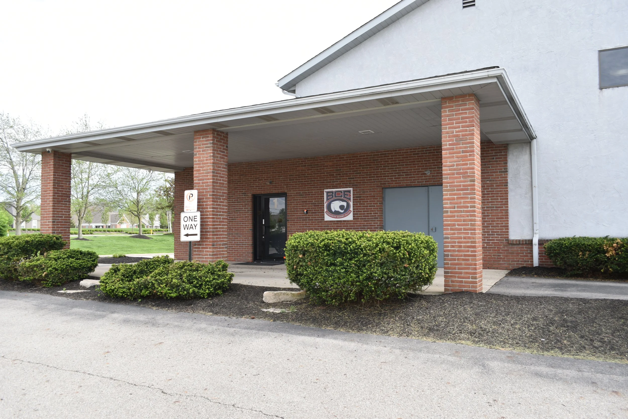 Exterior of a brick and white building with a covered entrance, surrounded by bushes and a parking lot, with a no parking sign and a logo with a bear and the letters BCE.