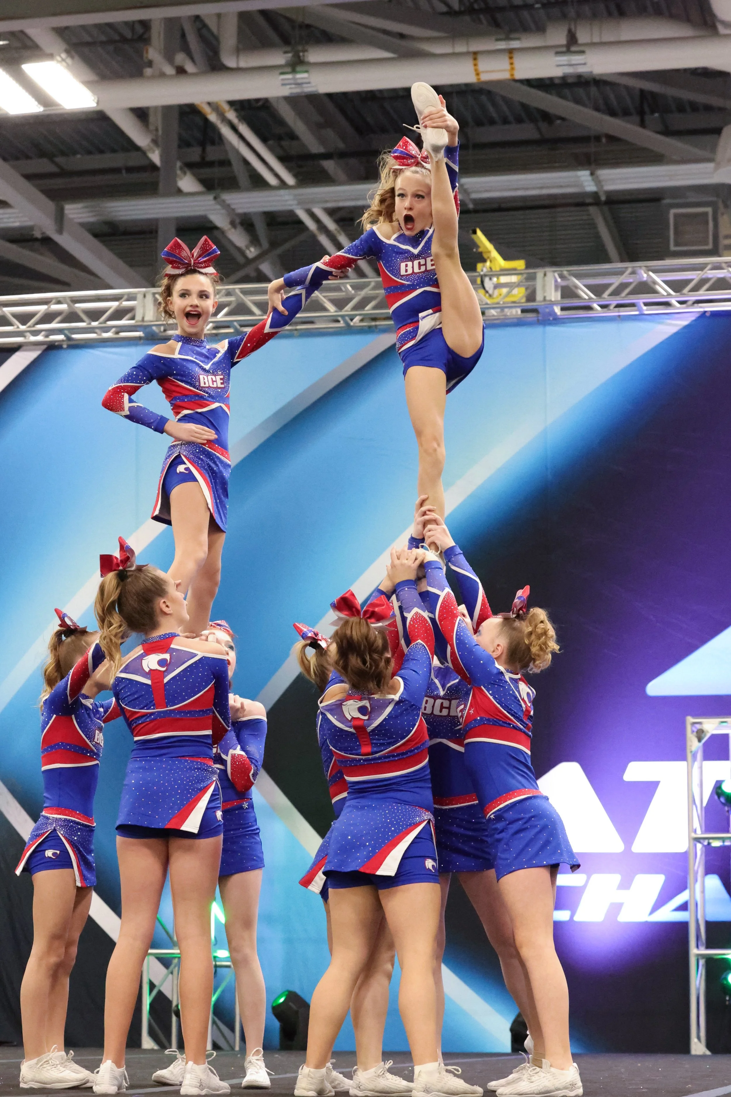 A group of young cheerleaders in blue, red, and white uniforms performing a cheer routine. Two cheerleaders are elevated above the others, with one balancing on her teammates' hands and the other standing on her hand with a high leg extension. They wear matching bows and cheer shoes. The background shows a stage with a blue and black backdrop and some light fixtures.