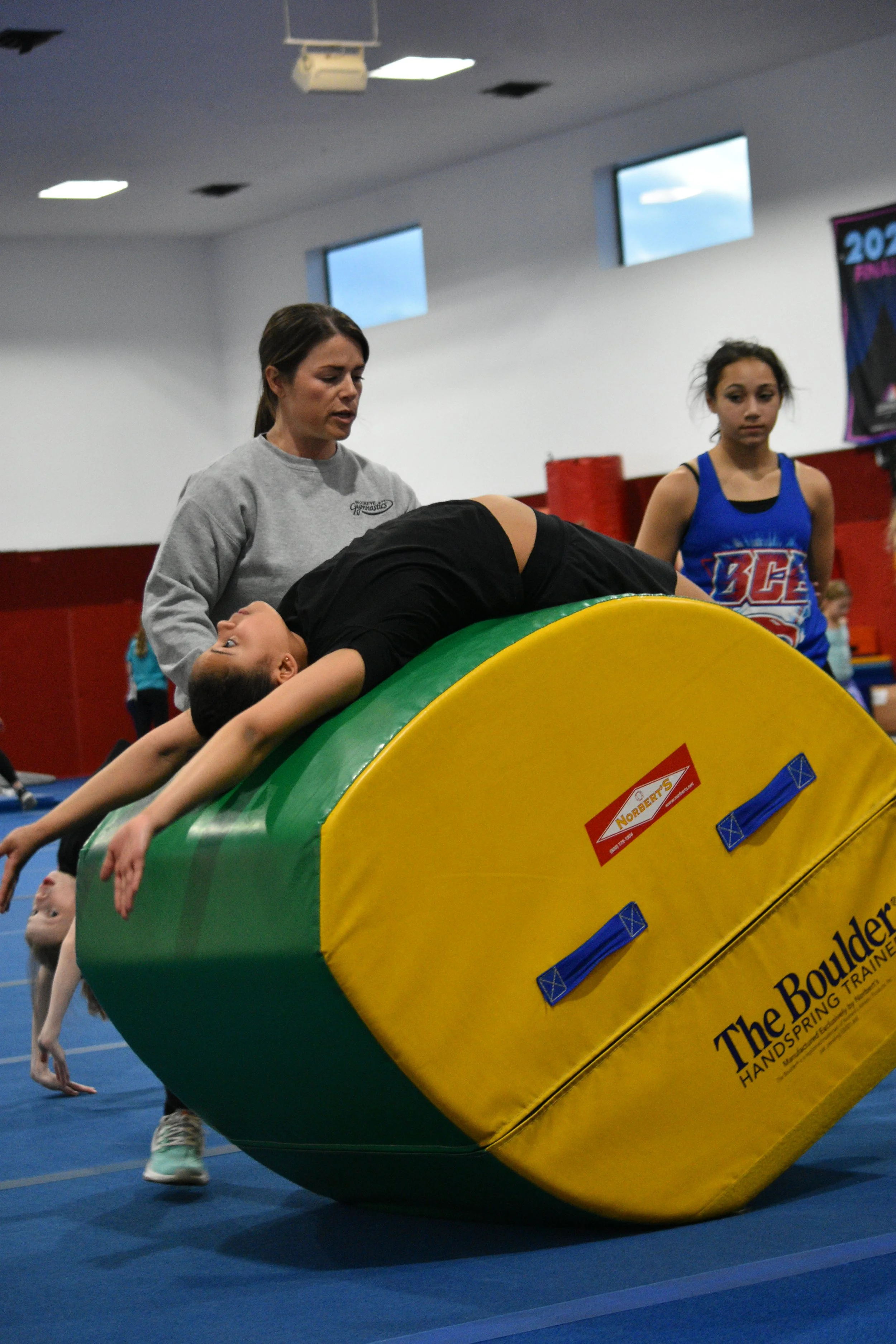 A girl in black sportswear is lying on a large, colorful gymnastics springboard inside a gymnasium, with a woman in a gray sweatshirt assisting her. Two other girls observe nearby, and a poster hangs on the wall in the background.