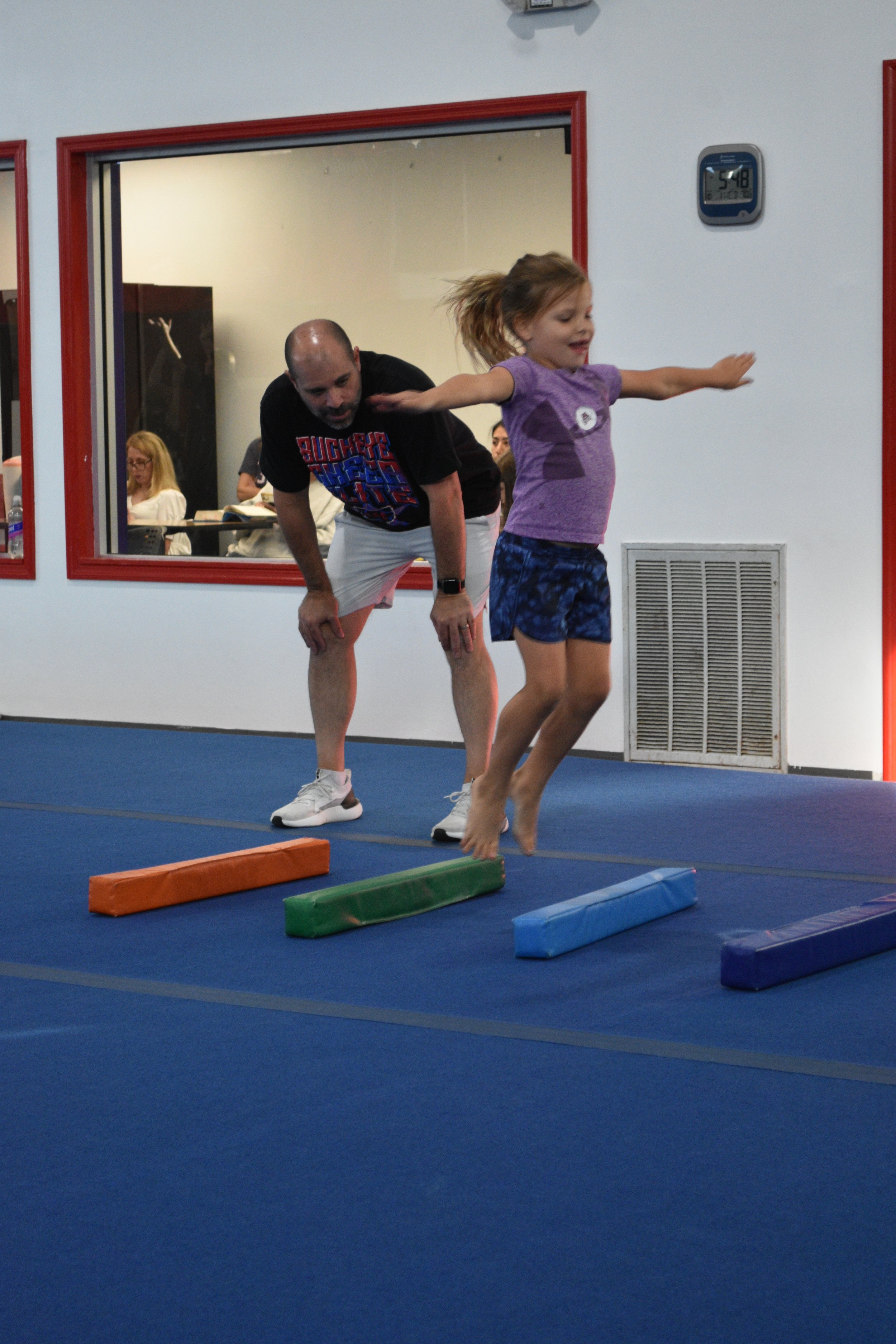 A young girl in a purple shirt and blue shorts practicing gymnastics with an instructor in an indoor gym. The girl is balancing on a foam block, arms extended, with colorful foam blocks on the floor in front of her. The instructor, dressed in a black t-shirt and shorts, is standing behind her, guiding her. Several people are seen through the window to another room, some seated at tables.