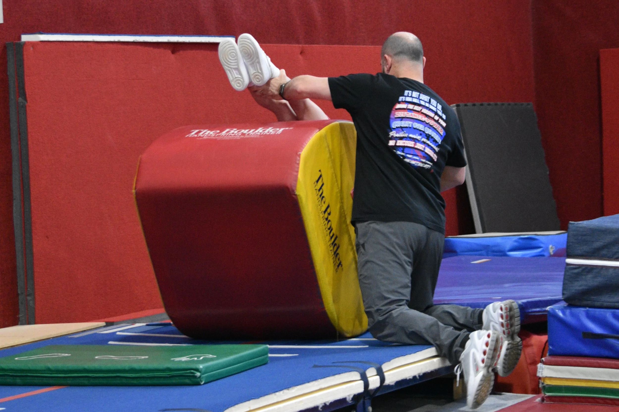 A man doing a handstand against a padded wall in a gym, with his feet pressed against the wall and his hands on the floor.