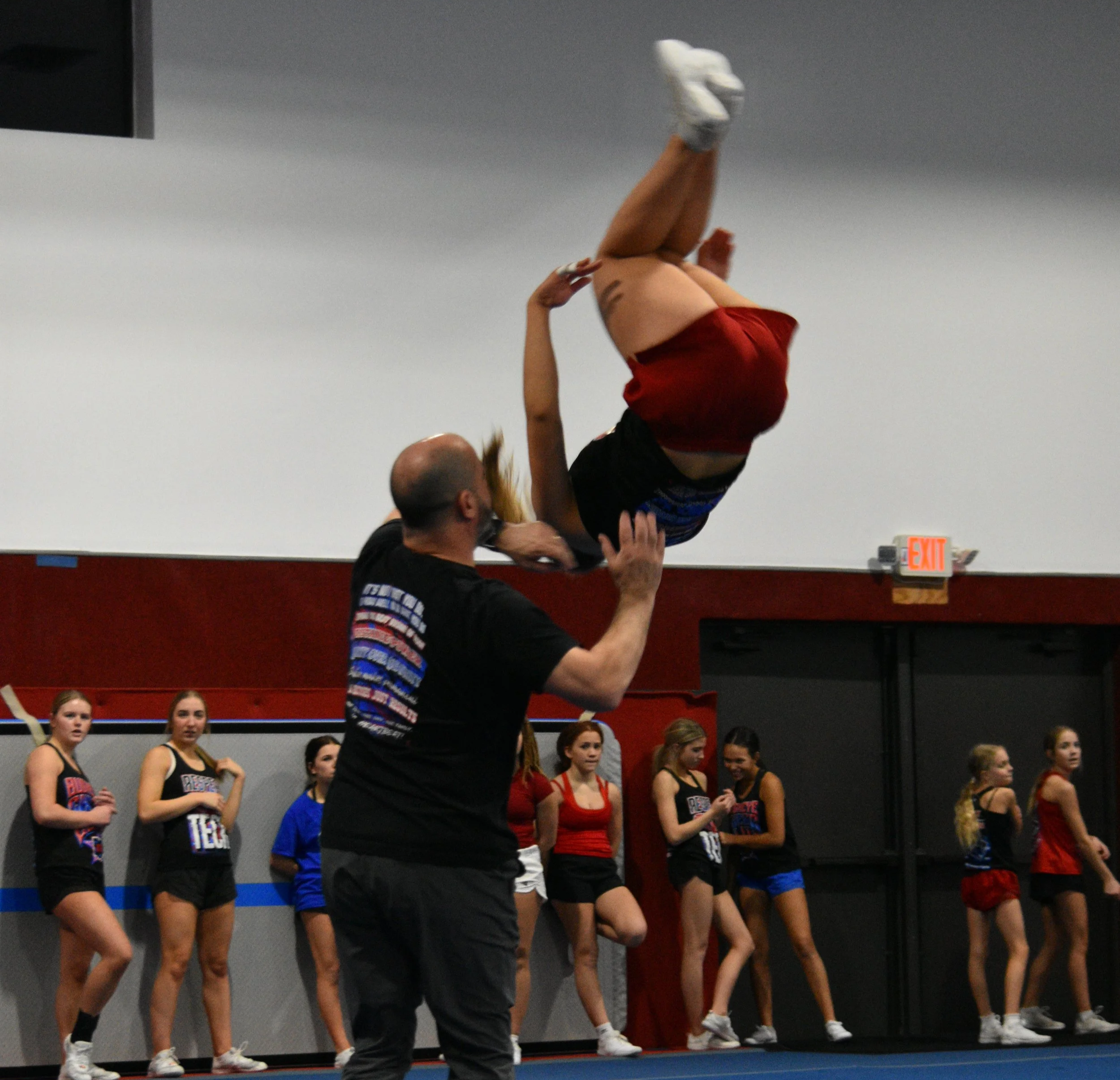 Cheerleaders standing in line during a cheerleading practice or event, with a person doing a stunt in the foreground.