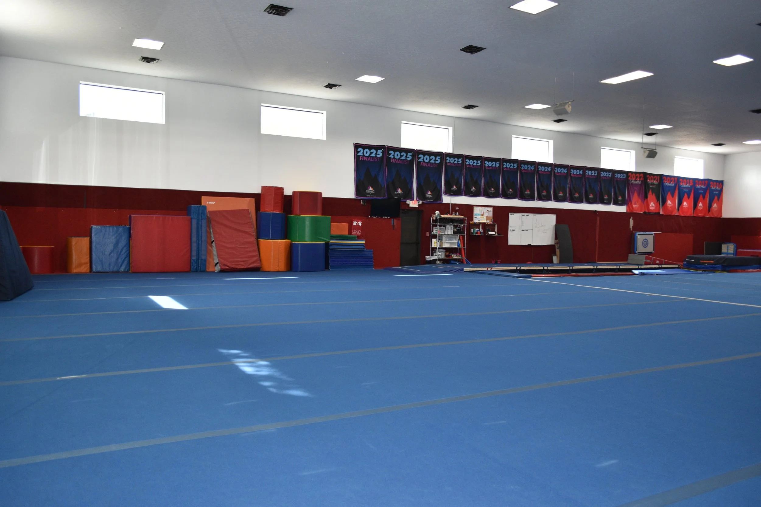 An indoor gym with blue mat flooring, various colorful padded equipment against the walls, and banners hanging from the ceiling displaying years from 2018 to 2025, indicating athletic events or competitions.
