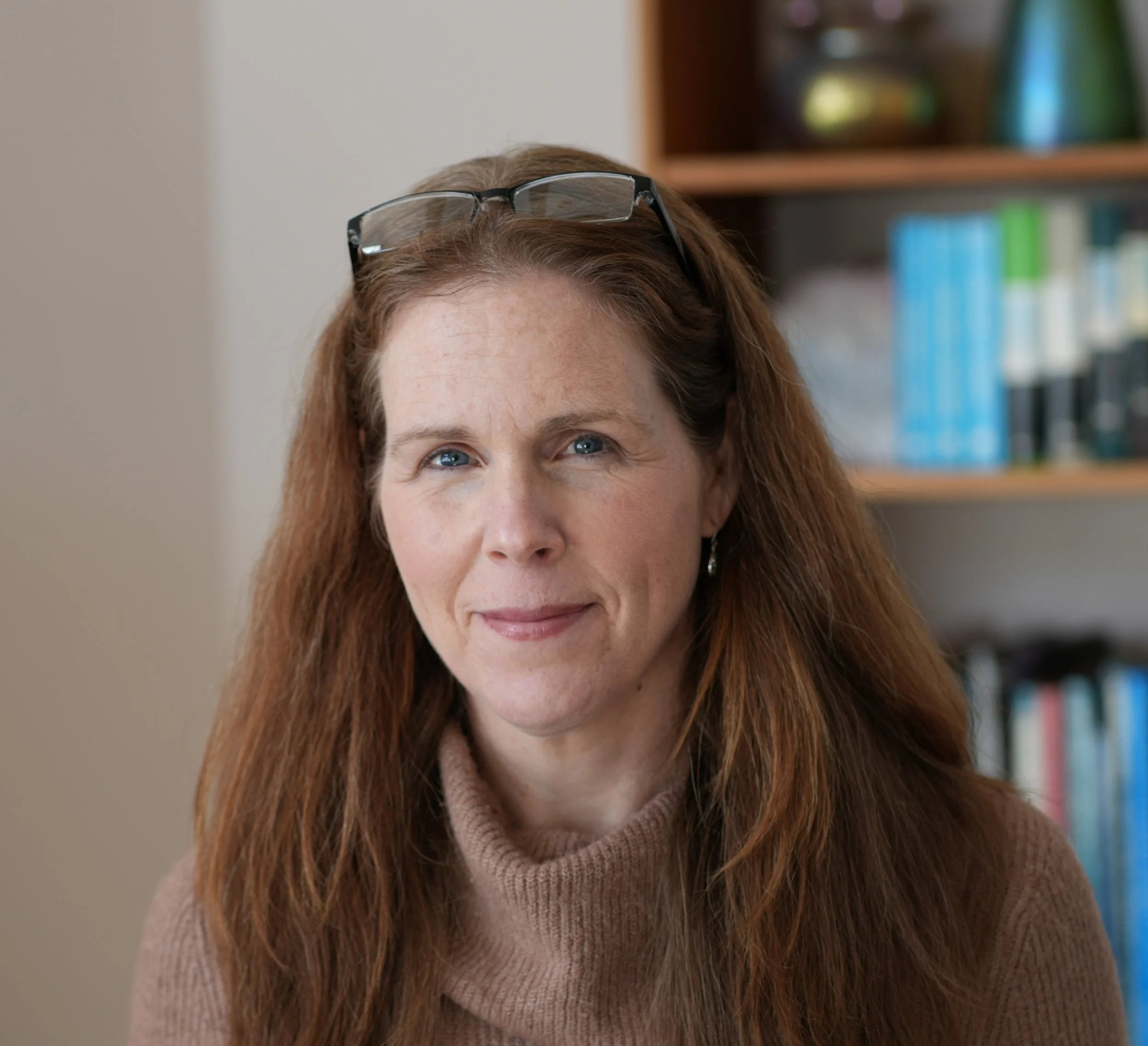Close-up of a middle-aged woman with red hair and blue eyes, wearing glasses on her head, indoors with a bookshelf in the background.