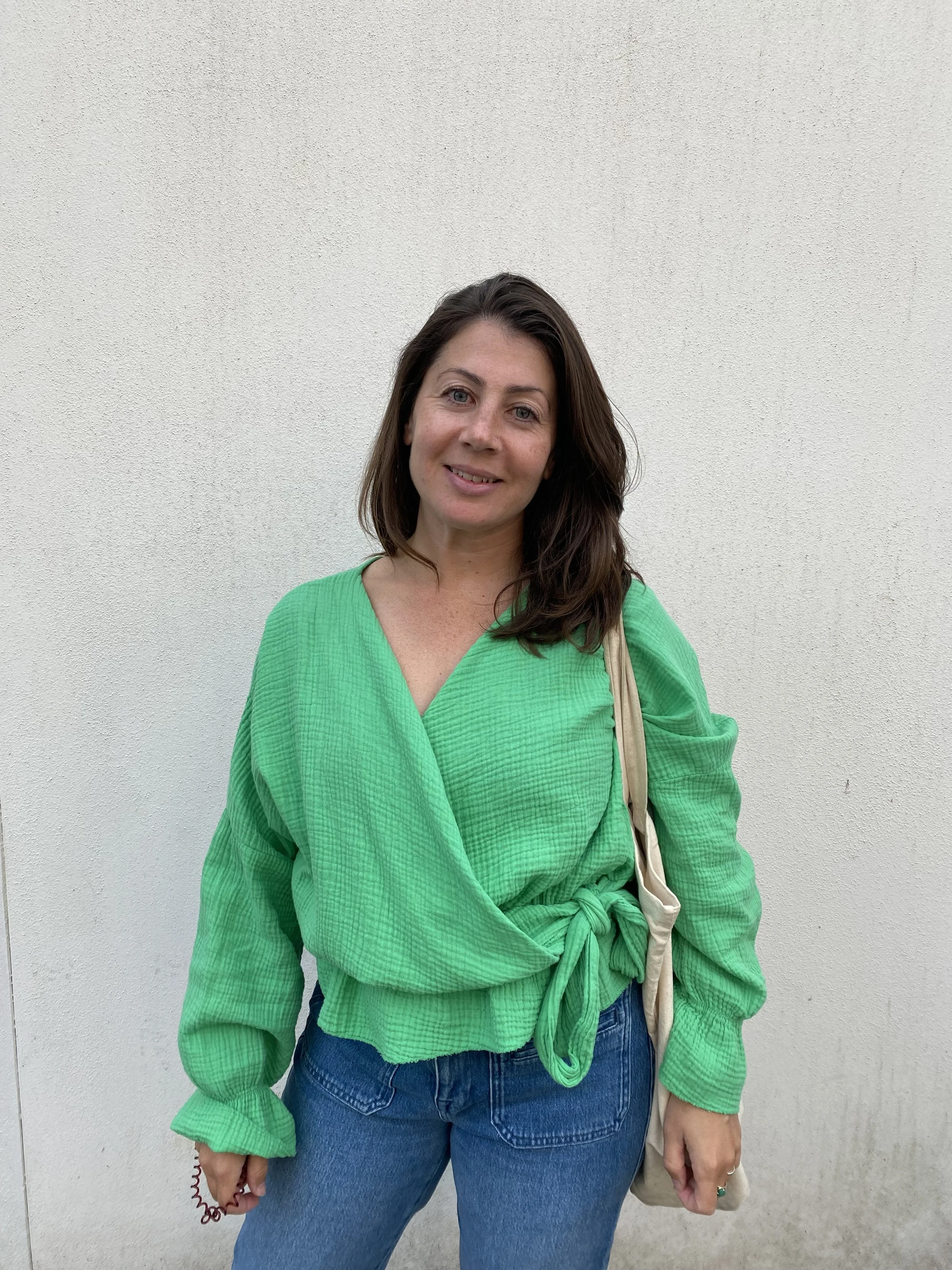 A woman with brown hair, wearing a bright green top with a waist tie, blue jeans, and carrying a beige tote bag, standing against a plain white wall.