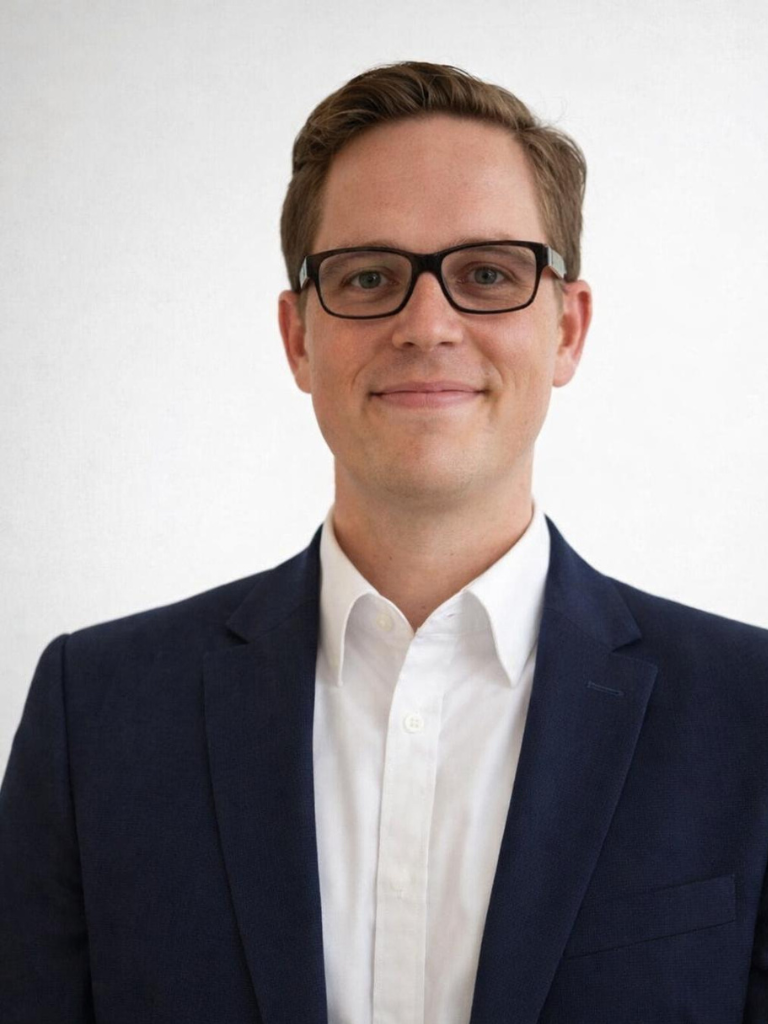 A man wearing glasses, a white dress shirt, and a dark blazer, smiling against a plain white background.