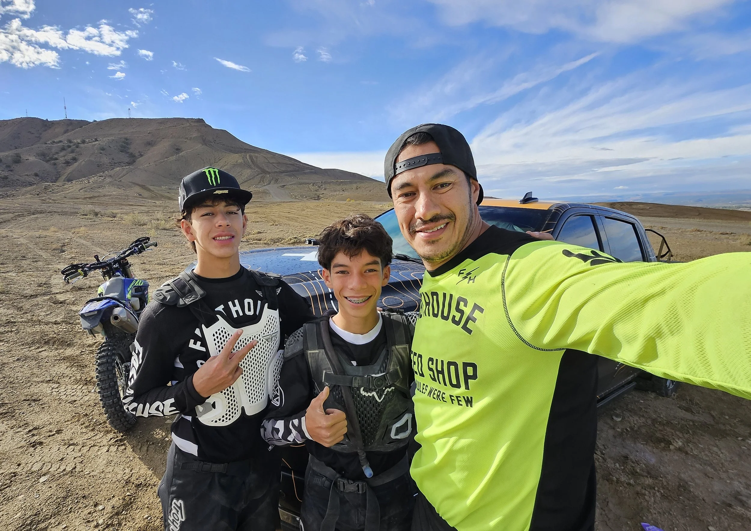 Three males taking a selfie outdoors near a black pickup truck with dirt terrain and mountains in the background. Two boys are wearing motocross gear, and an adult man is wearing a neon yellow shirt.