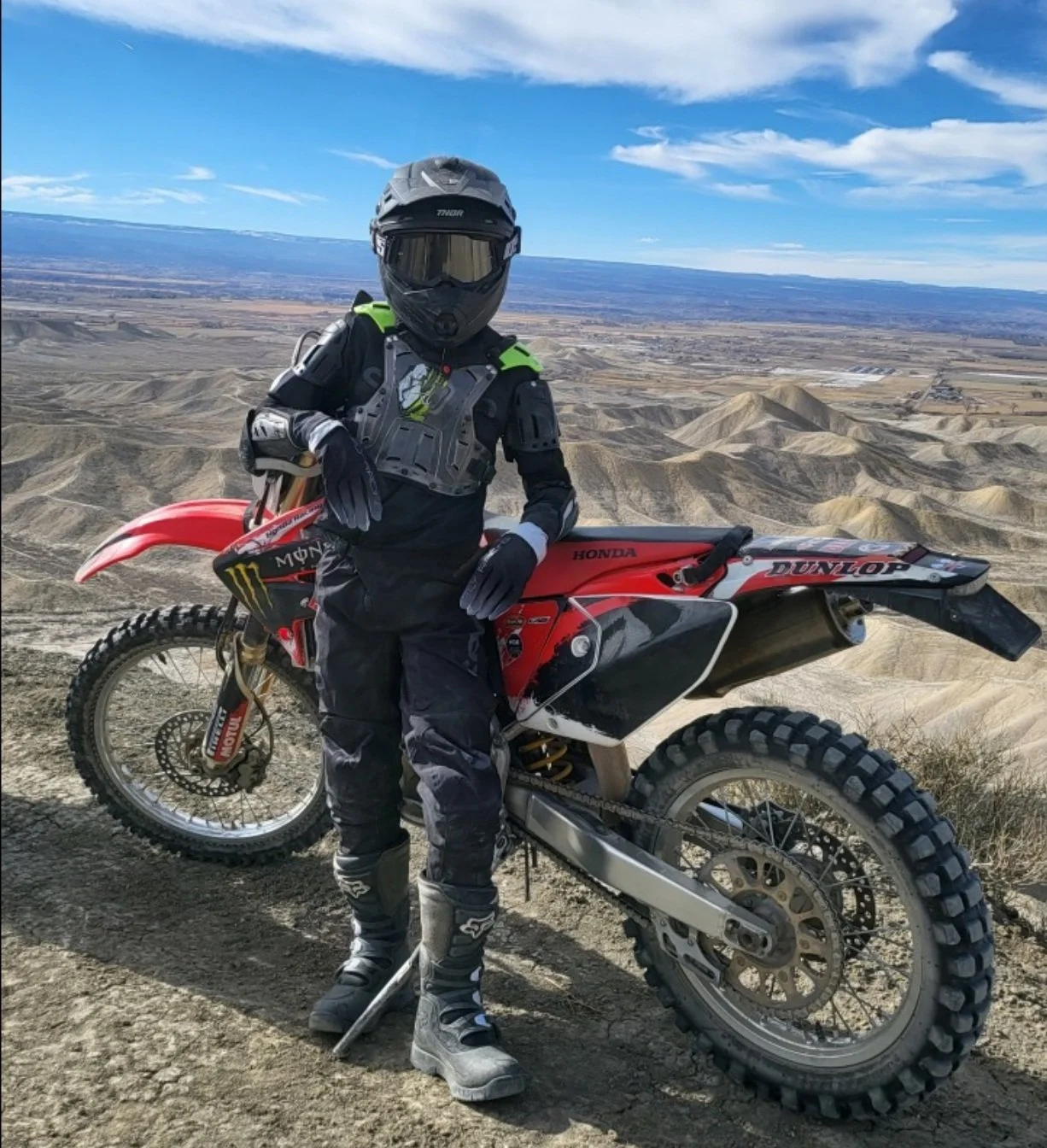 A person in motocross gear standing next to a red motocross bike on a dirt trail overlooking a desert landscape with rolling hills and a blue sky.