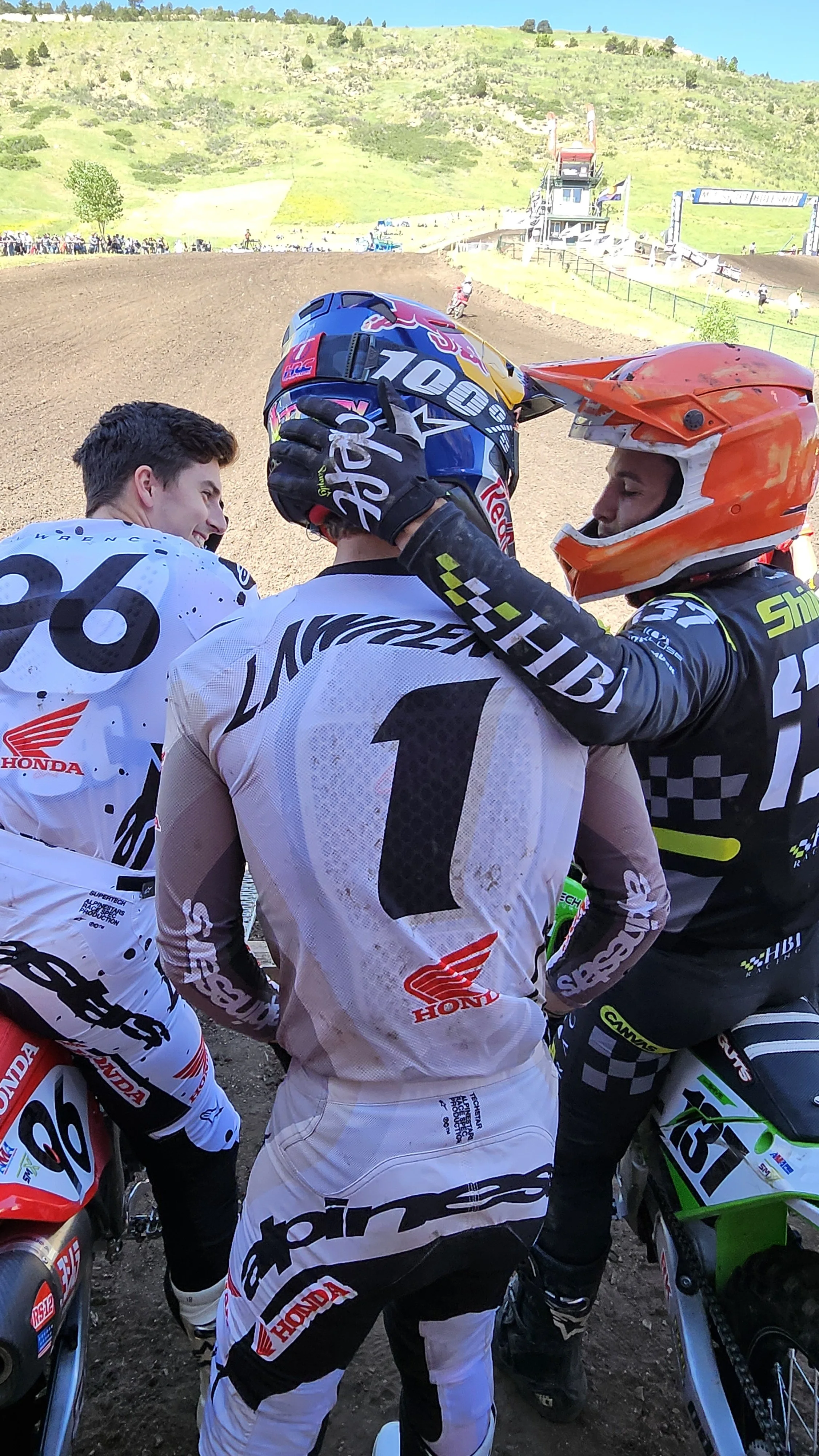 Motocross racers in a huddle before a race at a dirt track, with a hillside and spectators in the background.
