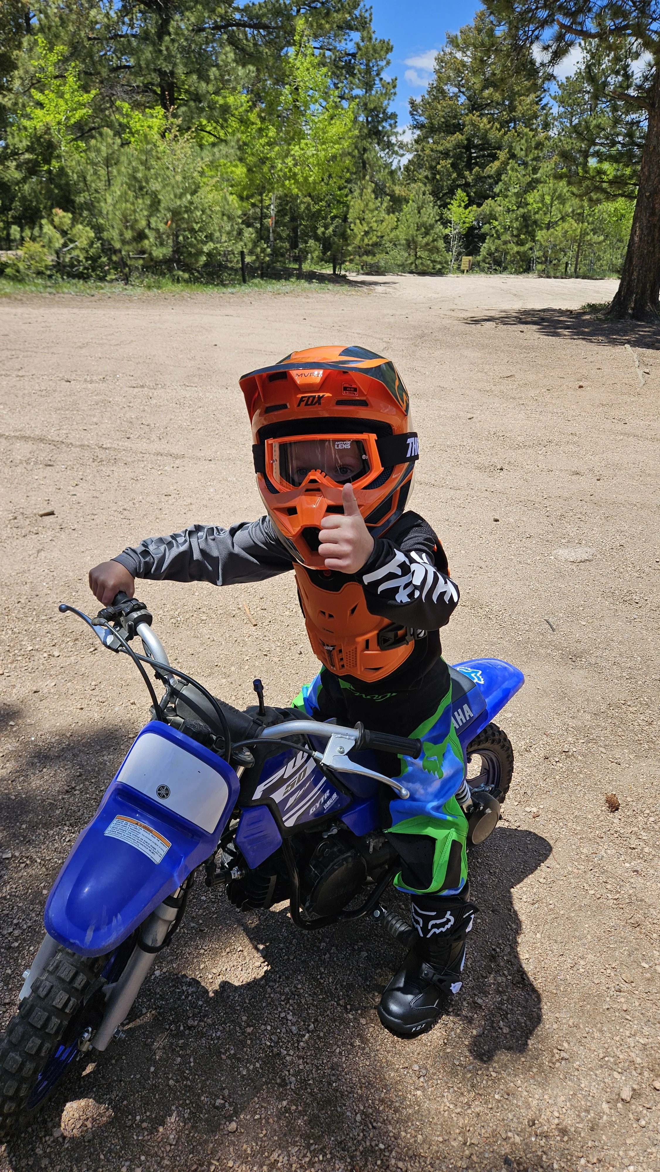 A young child dressed in motocross gear, wearing an orange helmet and goggles, giving a thumbs-up while sitting on a blue dirt bike in a wooded outdoor area.