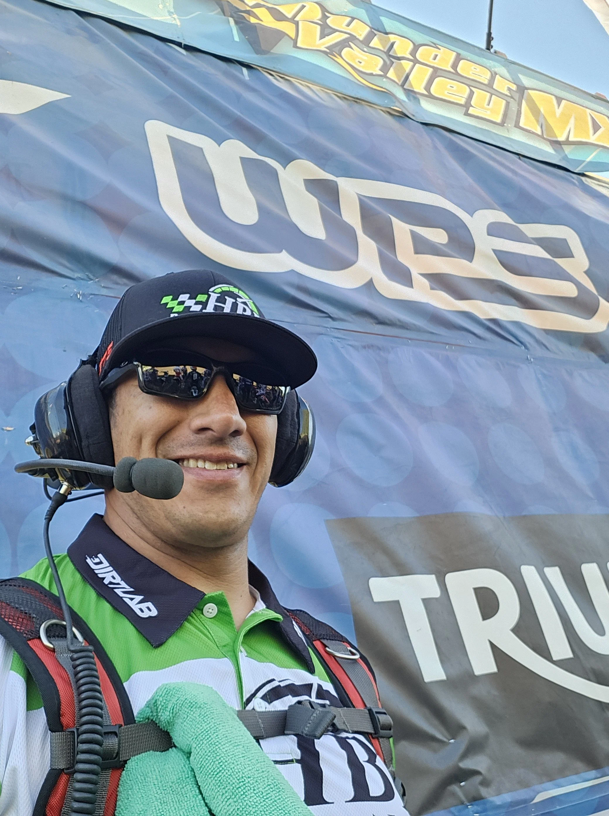 Smiling man in racing gear with headset and sunglasses in front of a promotional background at a racing event.