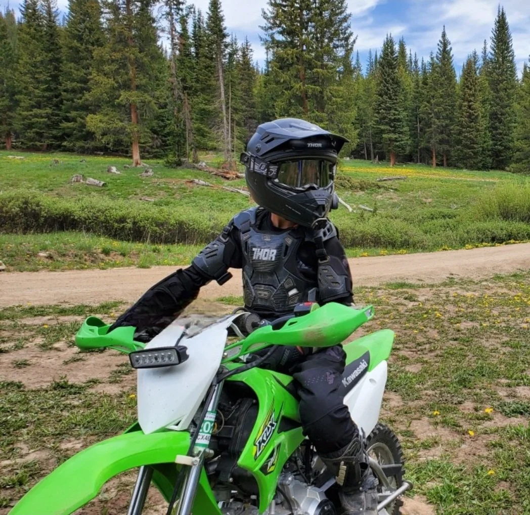 A child in black protective gear and a helmet sitting on a green Kawasaki dirt bike on a dirt path in a forested area with green trees and blue sky.
