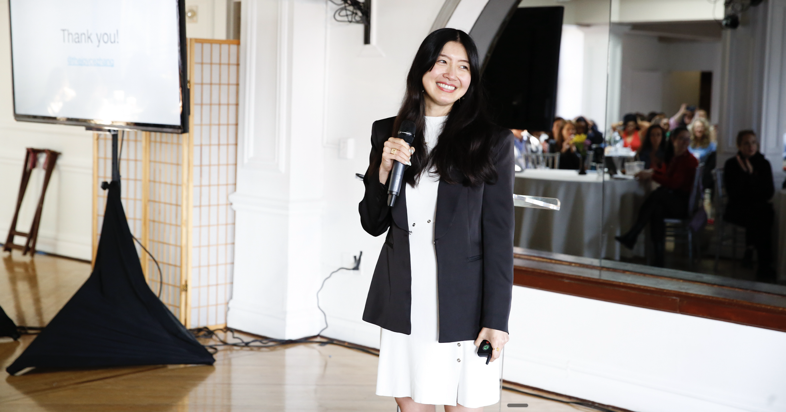 Joyce Zhang holding a microphone and smiling during a presentation at the Global Love Conference in NYC in May 2025 with an audience seated at tables in the background.