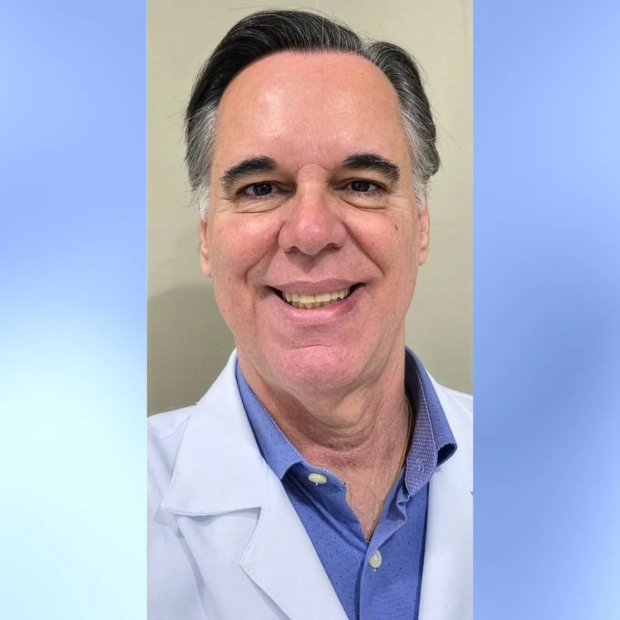 Homem sorridente com cabelo grisalho, vestindo jaleco branco e camisa azul, posando para foto de frente.