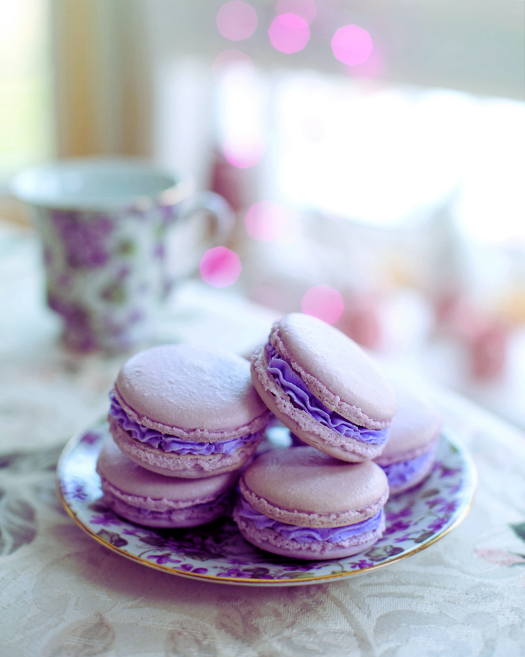 A plate of lavender macarons with purple filling on a table, with a floral teacup and a blurred background of pink lights.