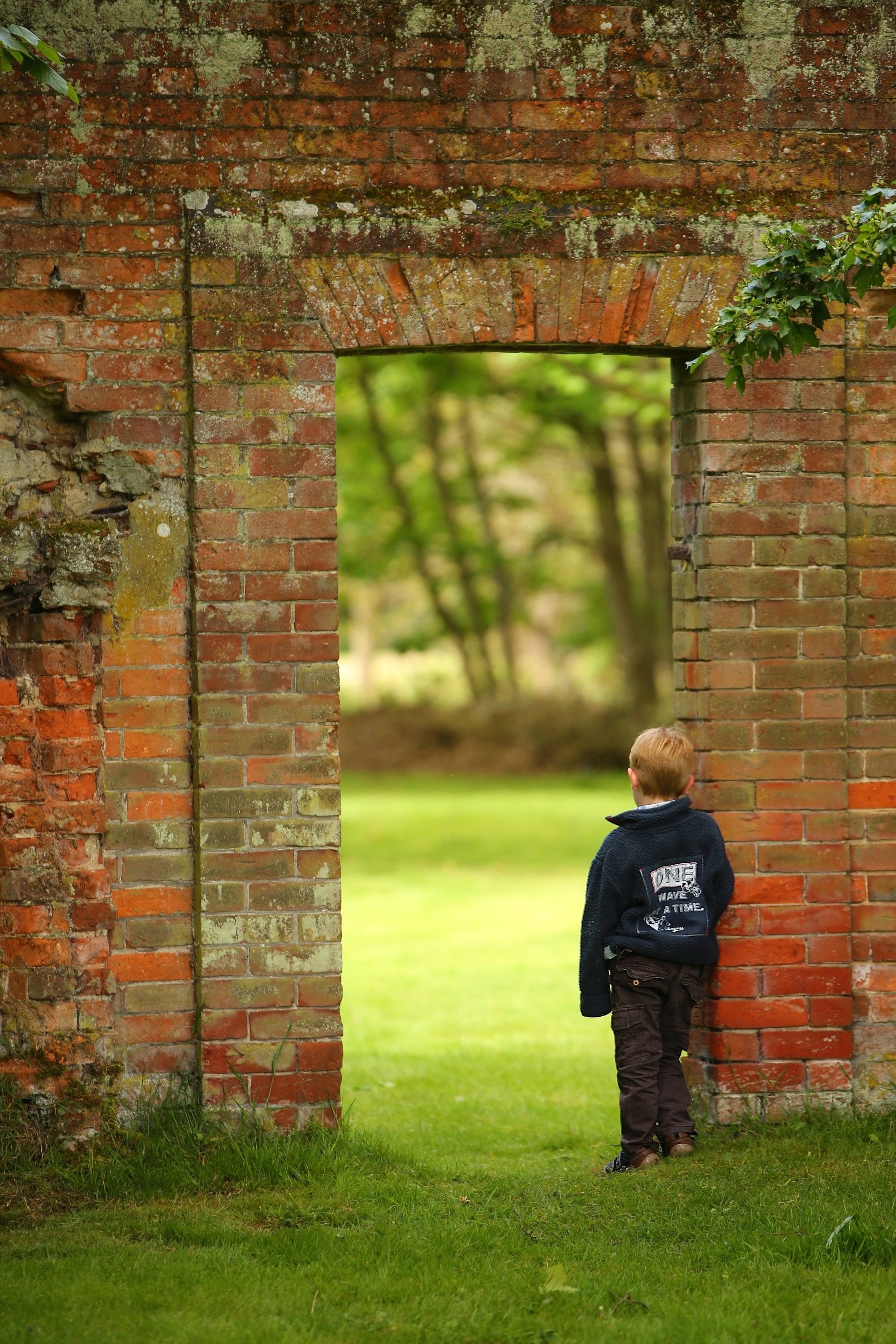 A boy with light hair dressed in dark clothing standing outside near a red brick structure with an opening, looking into a green wooded area.