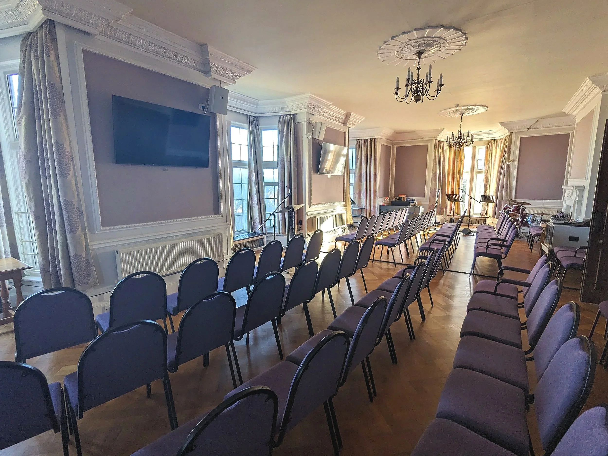 A spacious conference room set up for a presentation or event, with rows of chairs facing a small stage area, large windows with curtains, and ornate ceiling decorations with chandeliers.