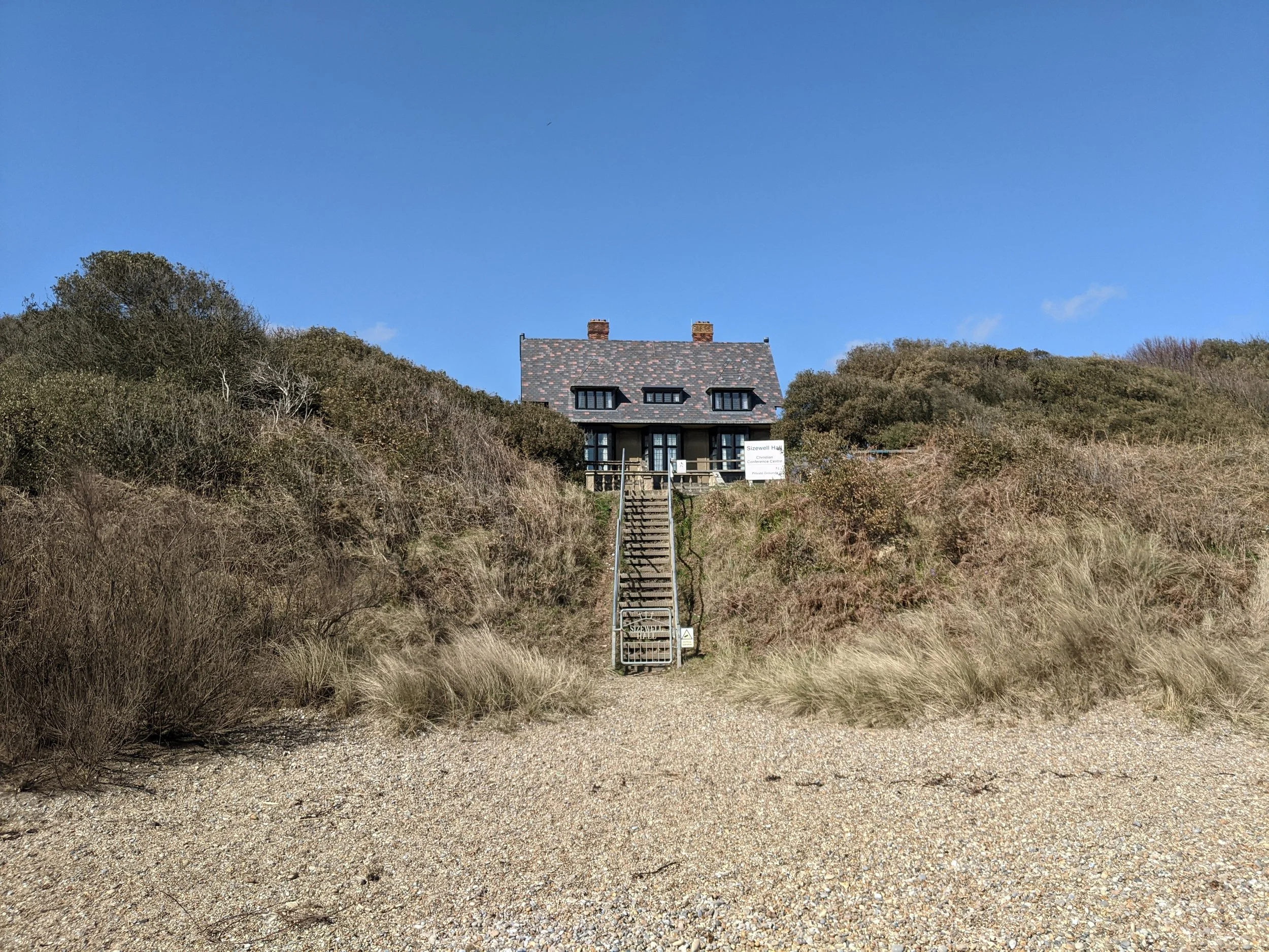 Sizewell Hall on the hill with a staircase leading up to it from the beach The house has a chimney and multiple windows and is surrounded by shrubbery and grass.