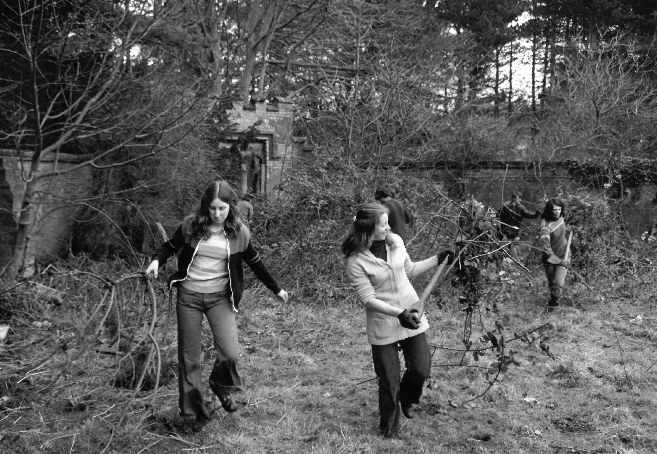 Black and white photo of four people walking through a wooded area with trees and bushes in the background.