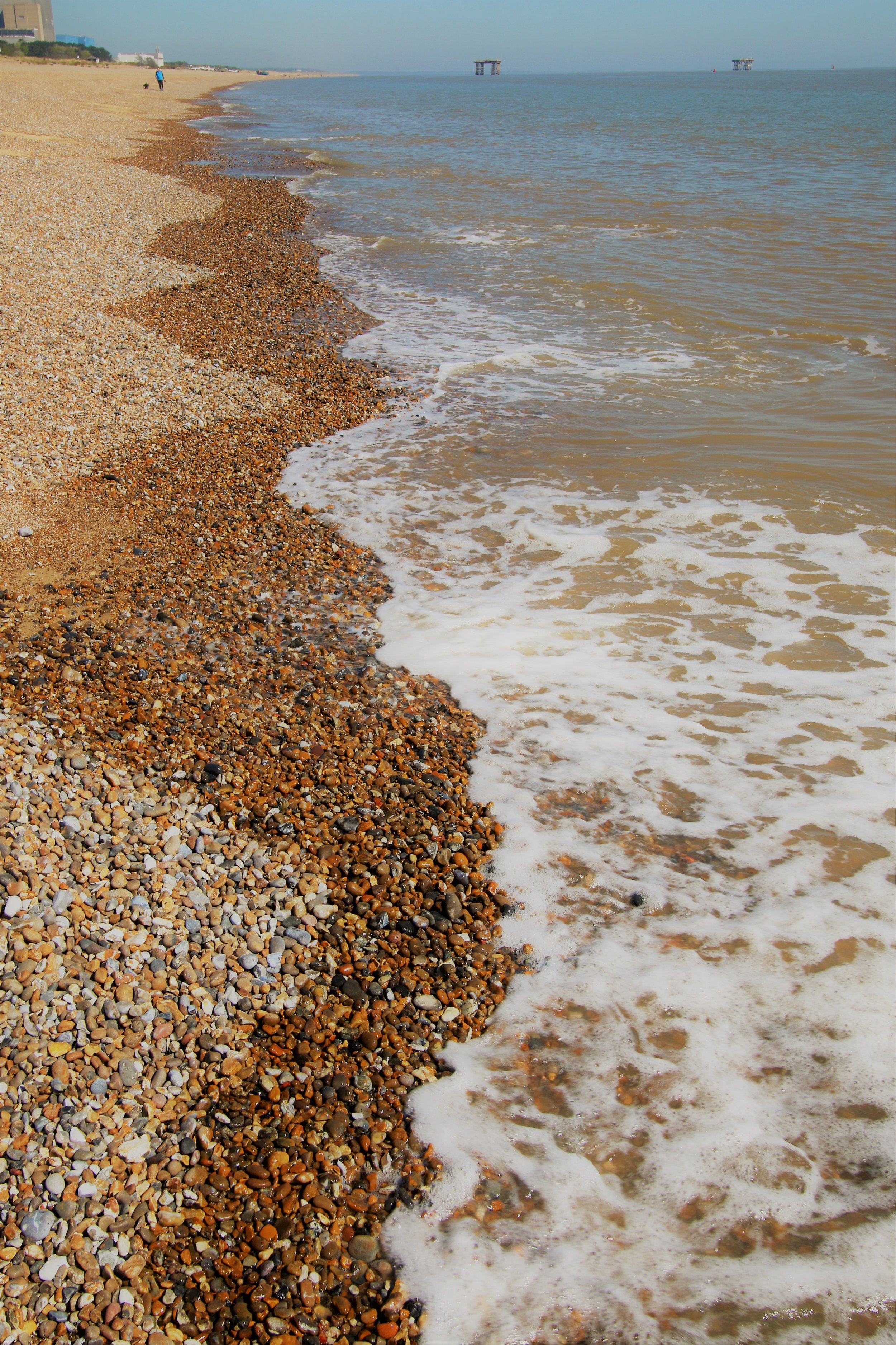 A beach with pebbles and small waves washing onto the shore, with a person walking a dog and in the distance some structures over the water.