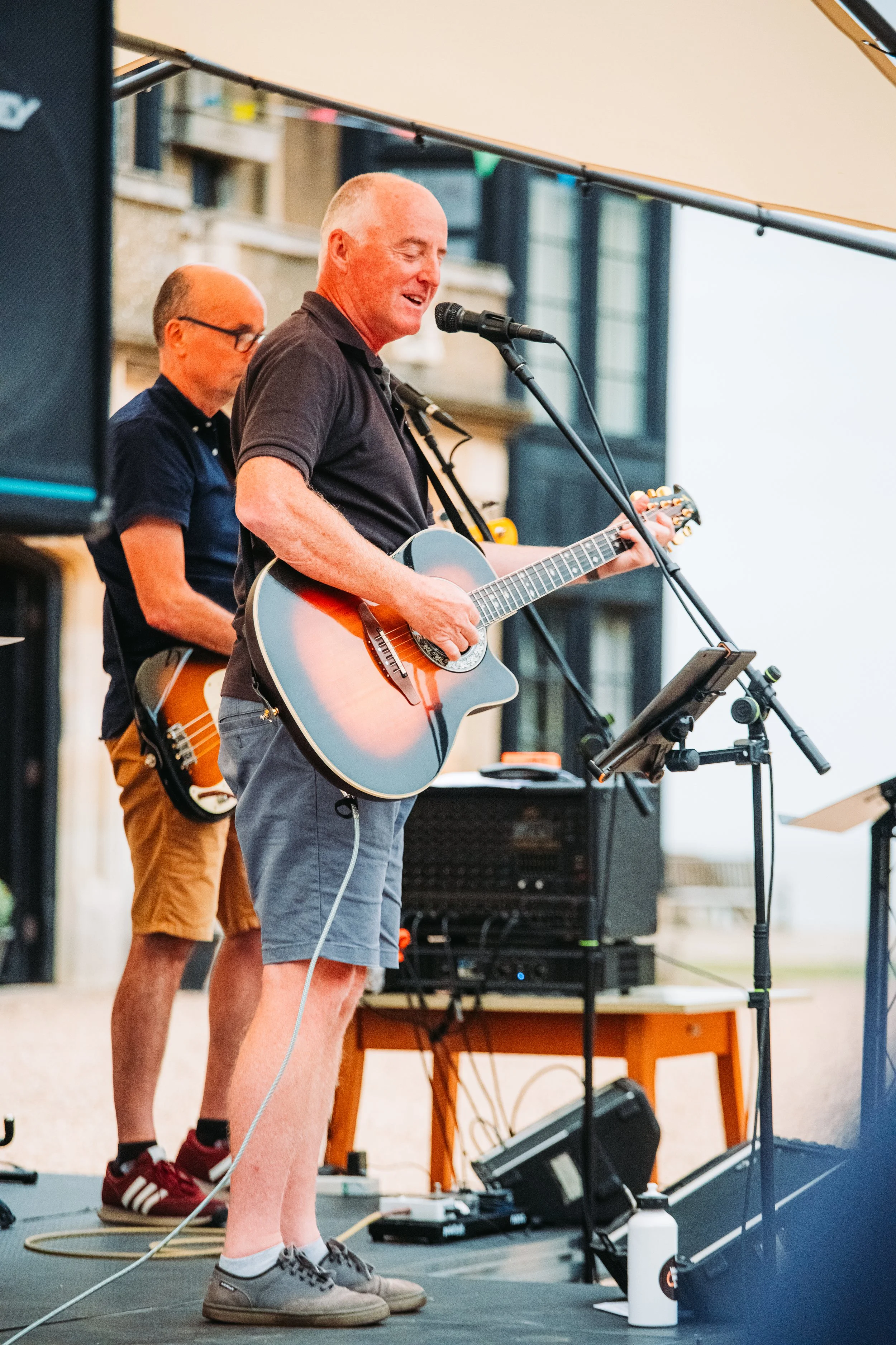 Tony hood playing an acoustic guitar and singing into a microphone at our 50th anniversary event with another person in the background also with a guitar, standing on a stage with equipment and a wooden table.