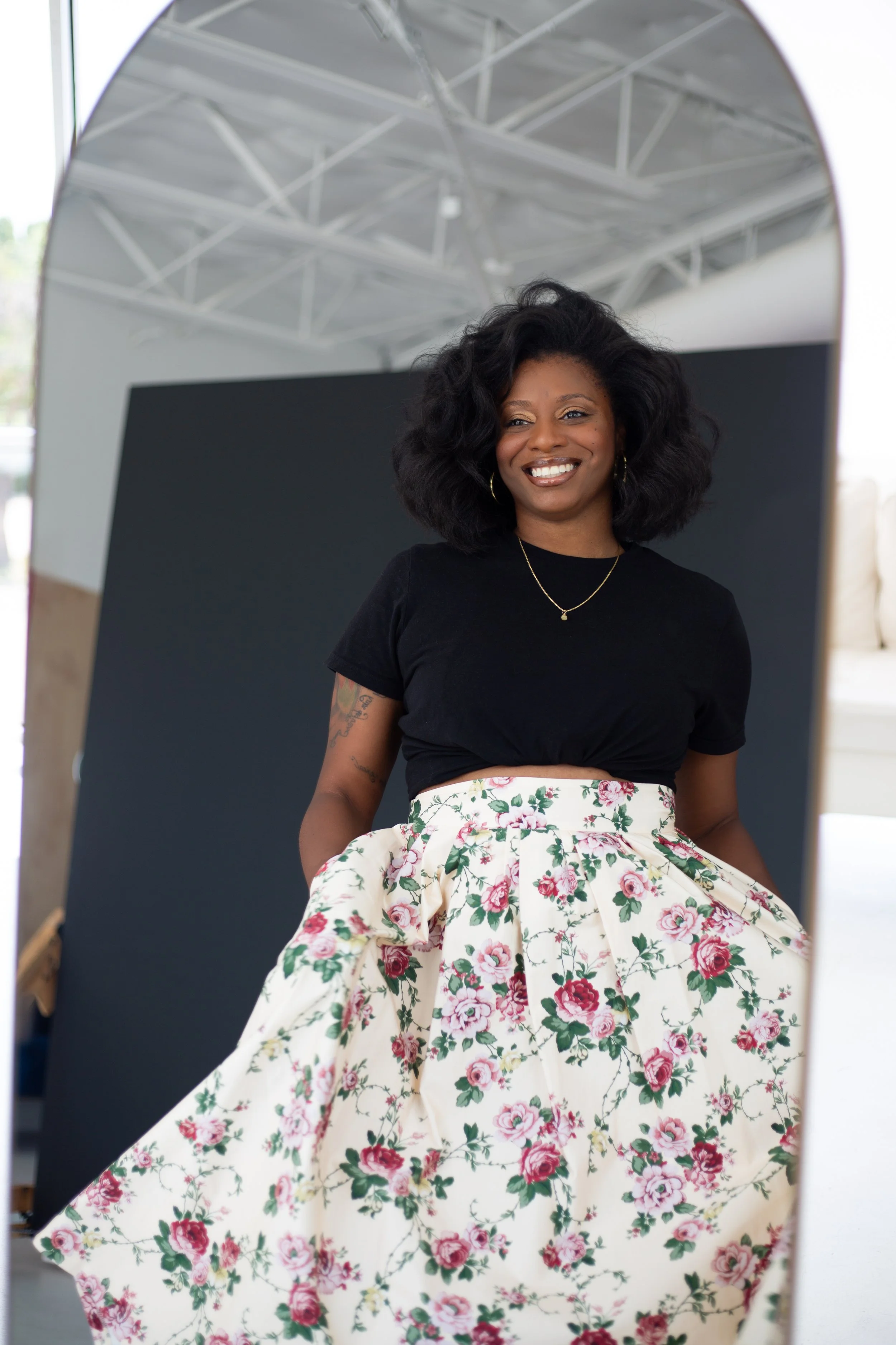 A woman with curly black hair, wearing a black top and a floral skirt, smiling as she looks into a mirror in a brightly lit room.