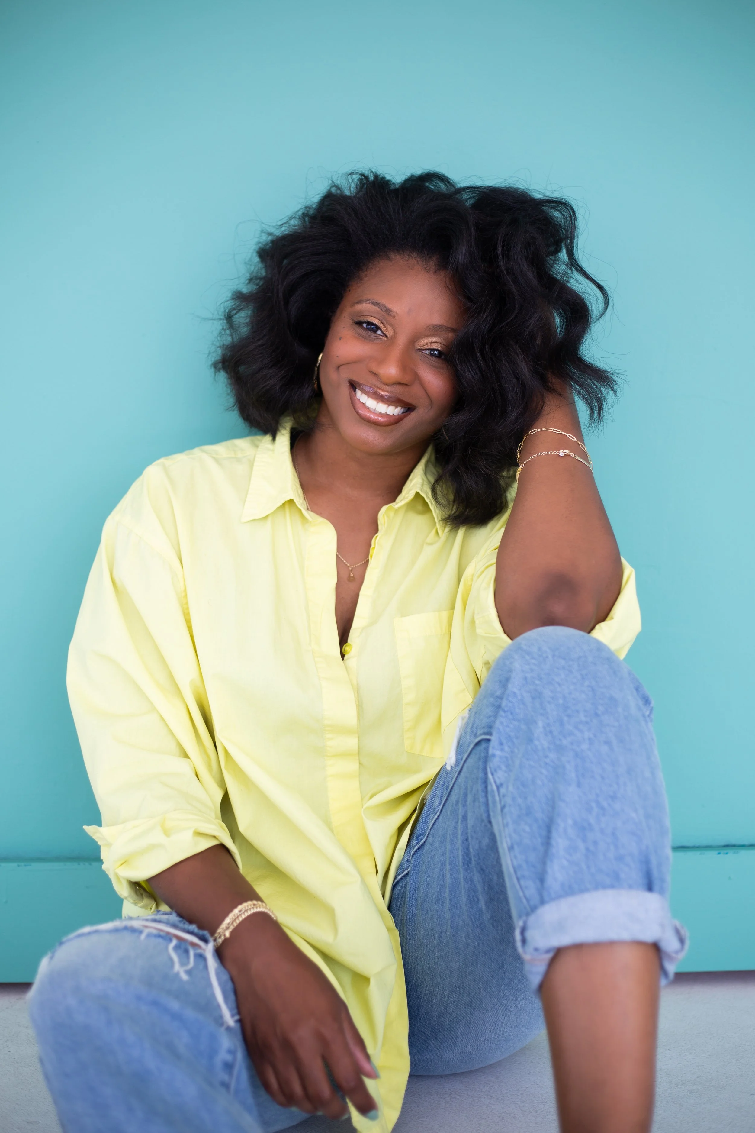 A woman with dark, curly hair smiling, wearing a light yellow shirt and ripped jeans, sitting against a turquoise wall.