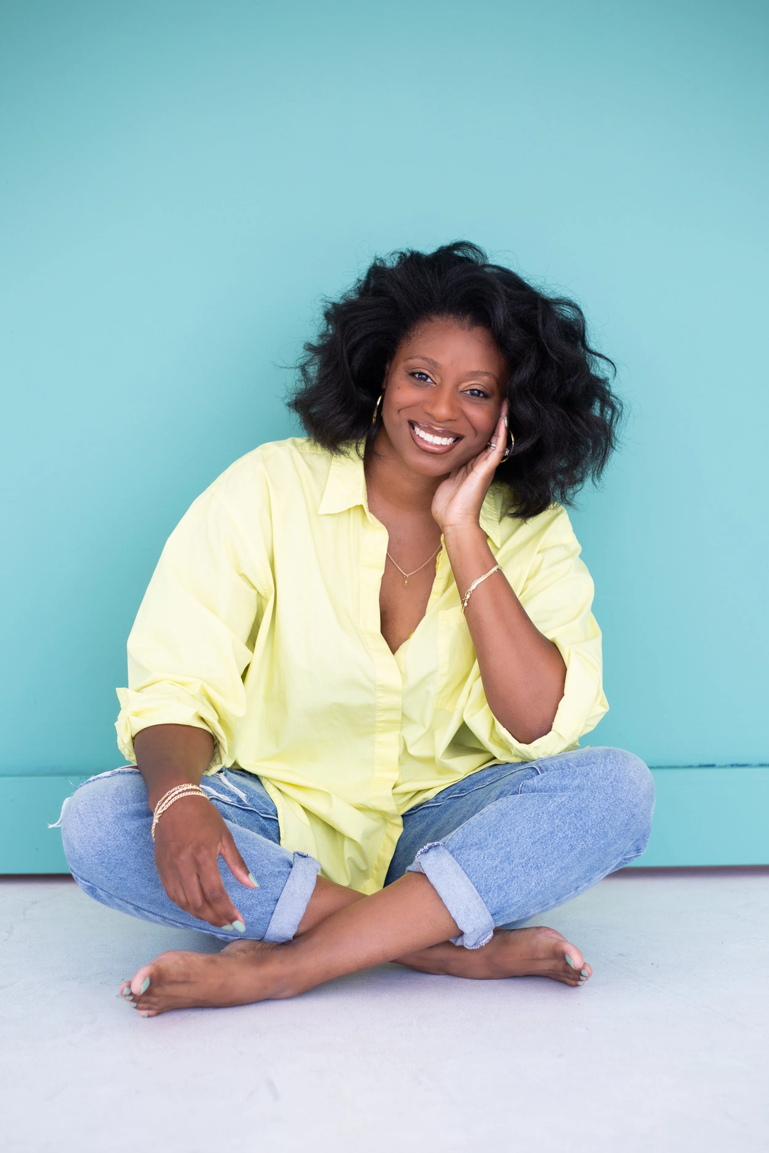A woman with black hair, wearing a yellow shirt and ripped jeans, sitting cross-legged against a turquoise wall, smiling and touching her face with her left hand.