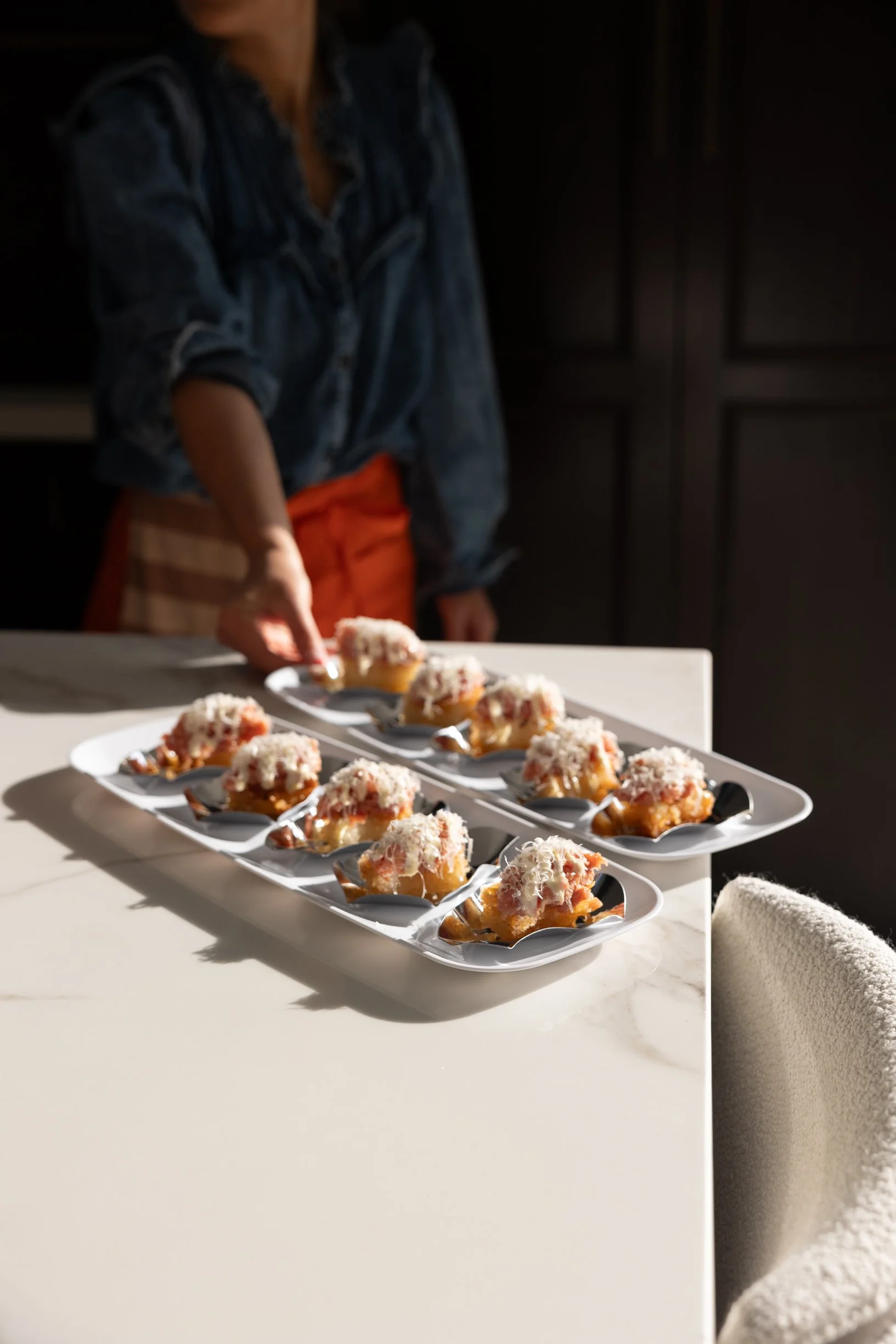 A person in a denim shirt prepares cupcakes with white frosting and shredded topping, arranged on a white serving platter on a white countertop.