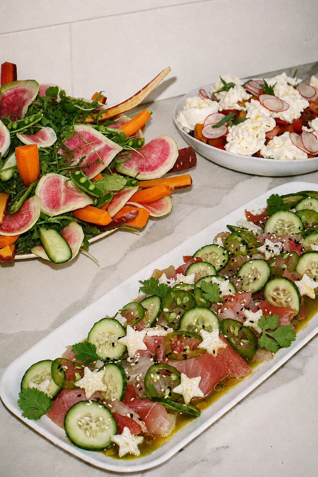 Various colorful salads on white plates and a large platter, including a salad with thinly sliced watermelon radish, carrots, and greens, a dish with crumbled cheese, cherry tomatoes, and radish slices, and a plate with cucumber, tomato, cilantro, and sesame seeds.