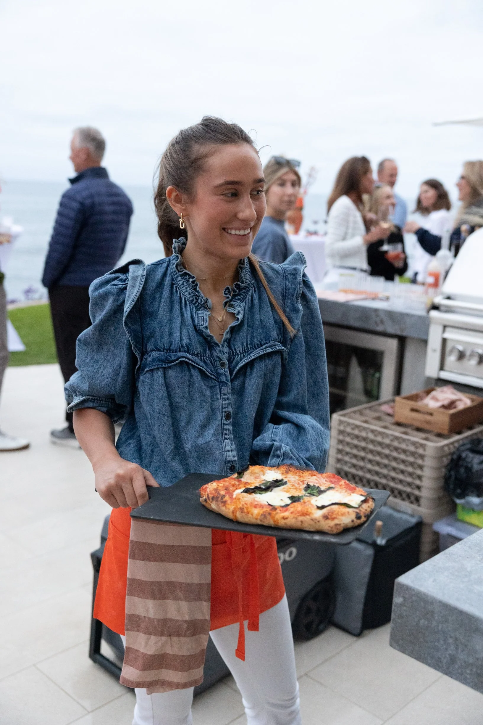 A young woman in a denim jacket and orange apron holds a tray with a freshly baked pizza topped with cheese, basil, and tomato at a social gathering near the coast. In the background, other people are talking and socializing.
