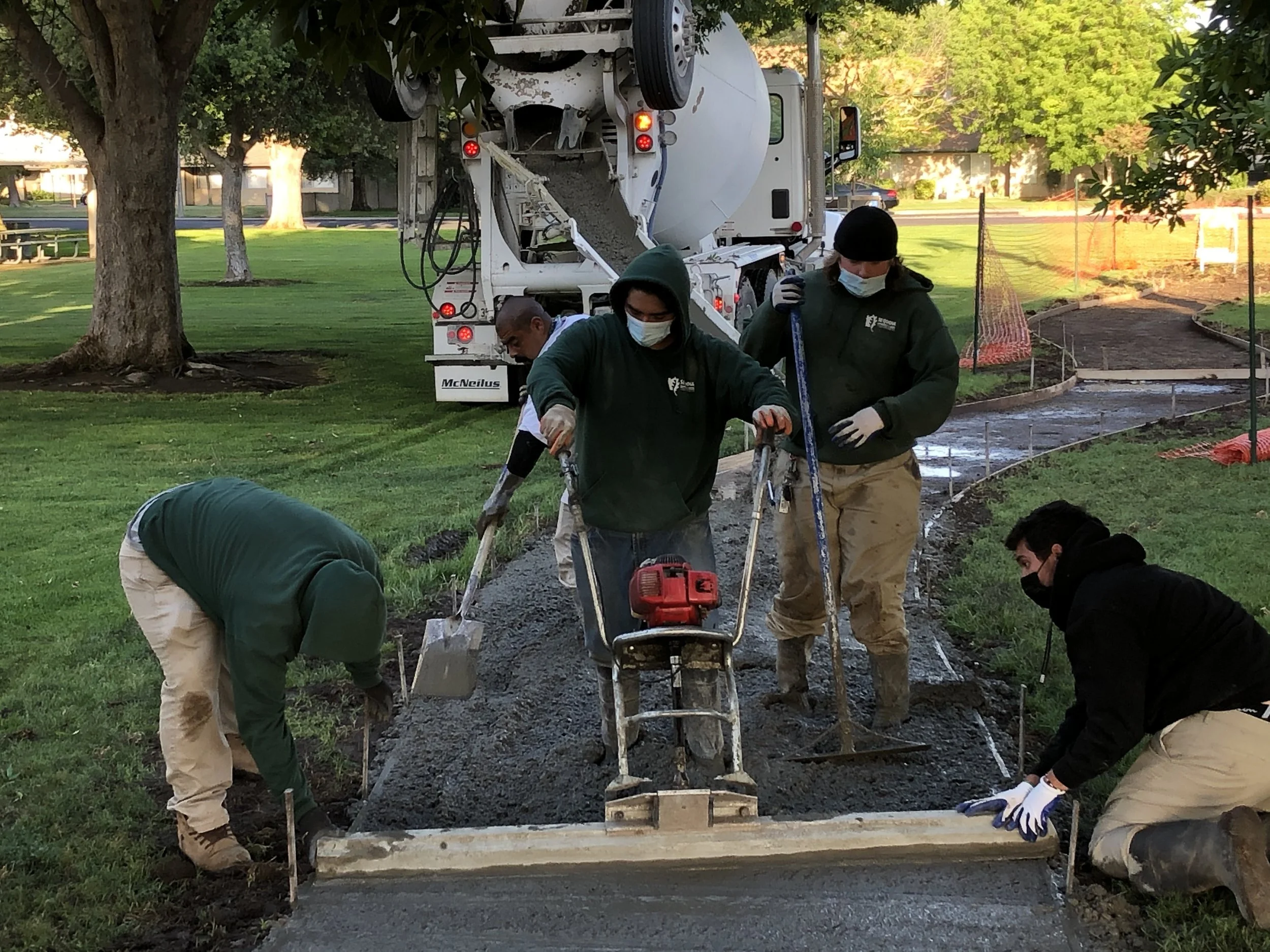 CSET workers in green sweatshirts and masks leveling wet concrete poured from a cement mixer for a sidewalk.