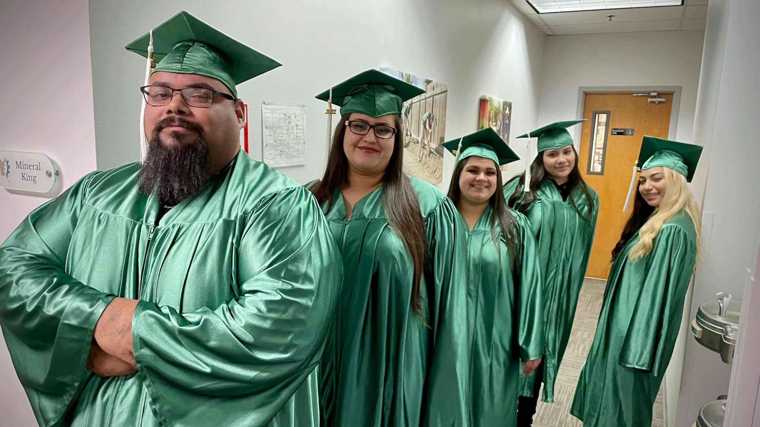 A group of smiling CSET adult graduates standing together in green caps and gowns inside a hallway.
