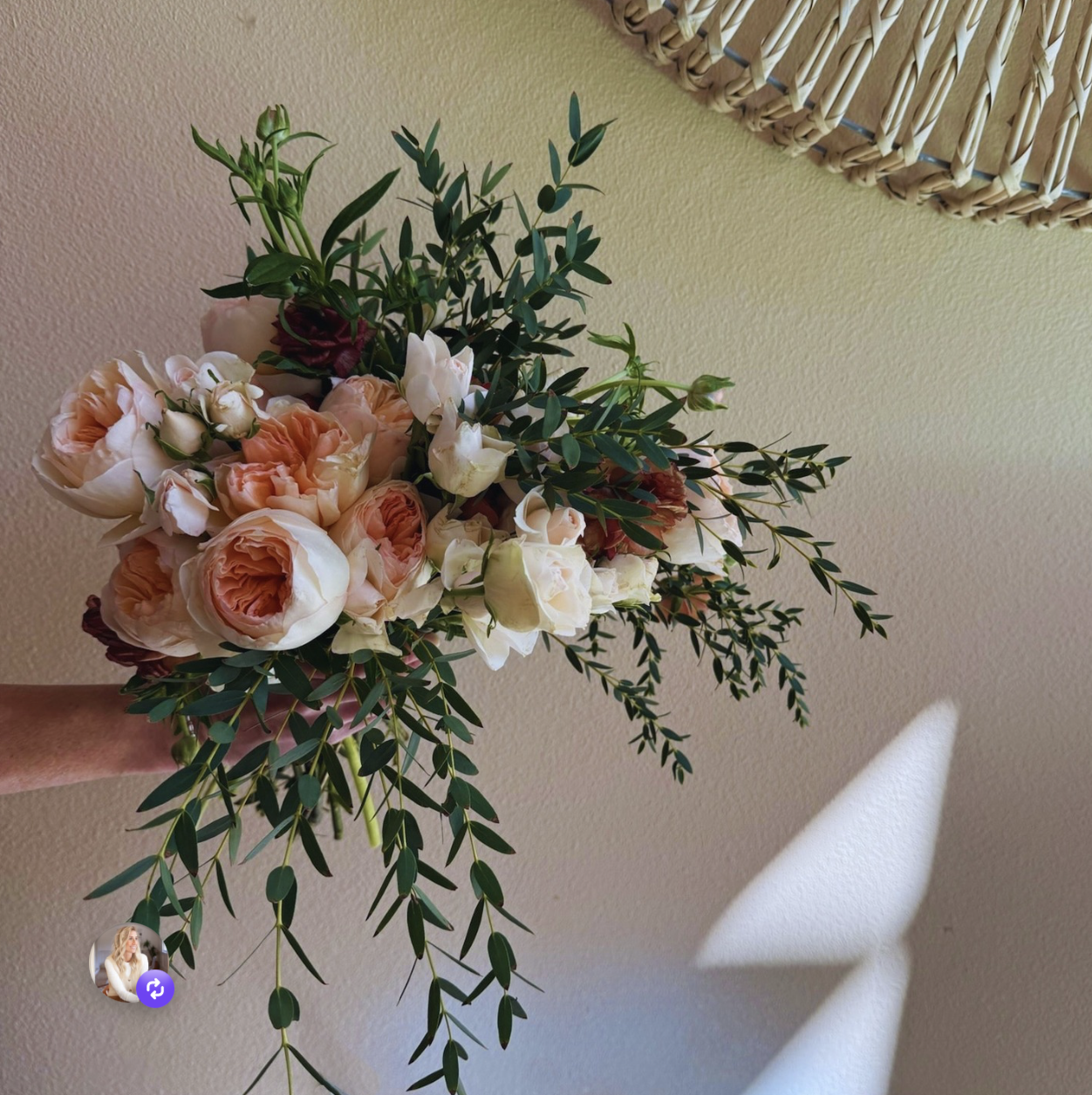Person holding a bouquet of peach and white roses with greenery in front of a beige wall and a wicker mirror.