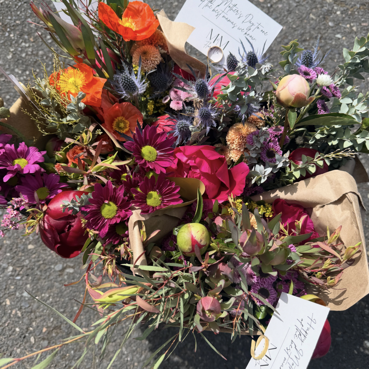 A colorful bouquet of mixed flowers including pink, red, purple, orange, and yellow blooms with green foliage, wrapped in brown paper, with a note and wedding rings attached.